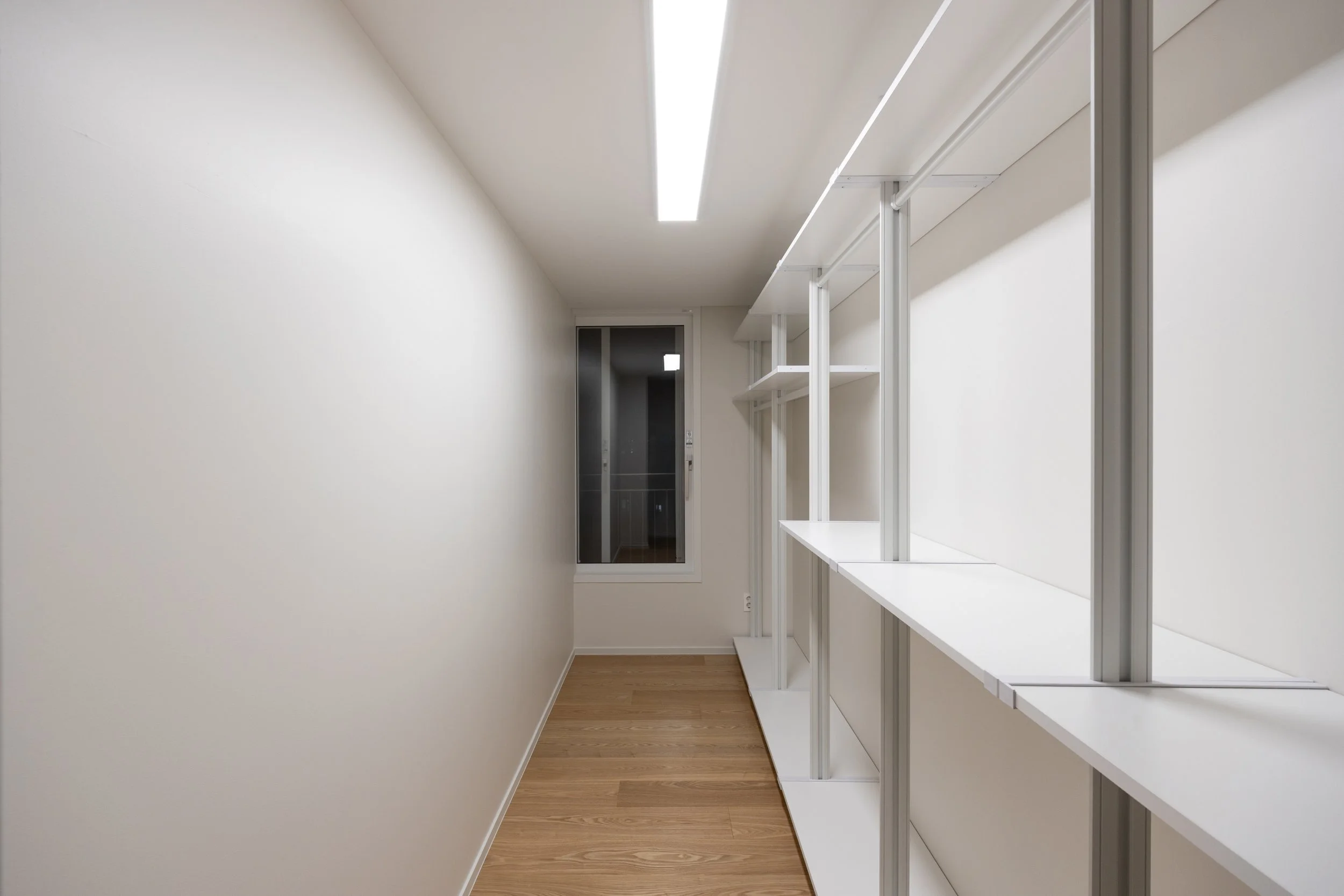 A narrow, empty room with white walls, a wooden floor, a ceiling light, a window at the end, and white shelving on the right side.