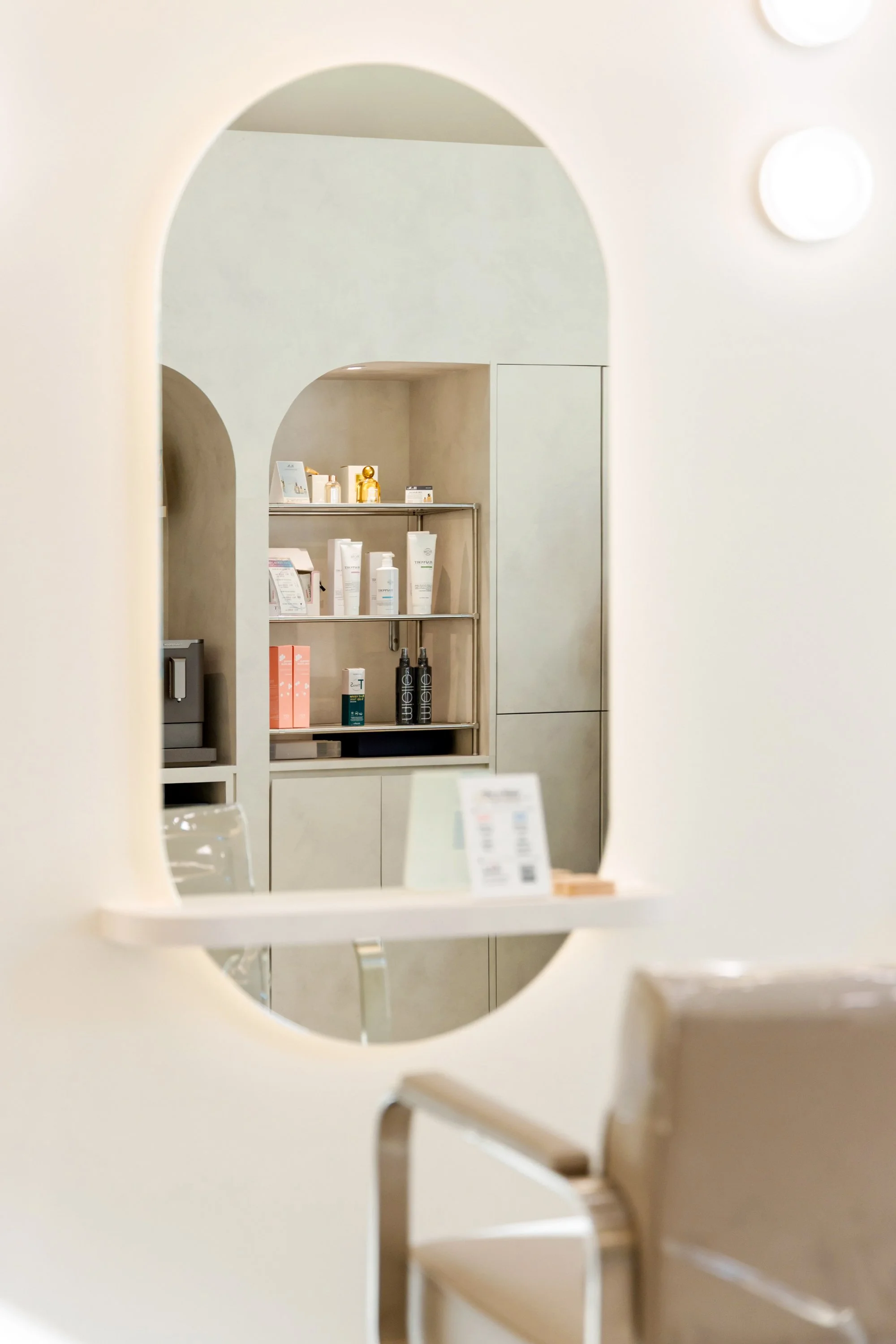 A view through a mirror showing a shelf with skincare products and a gold perfume bottle in a modern, minimalist interior.