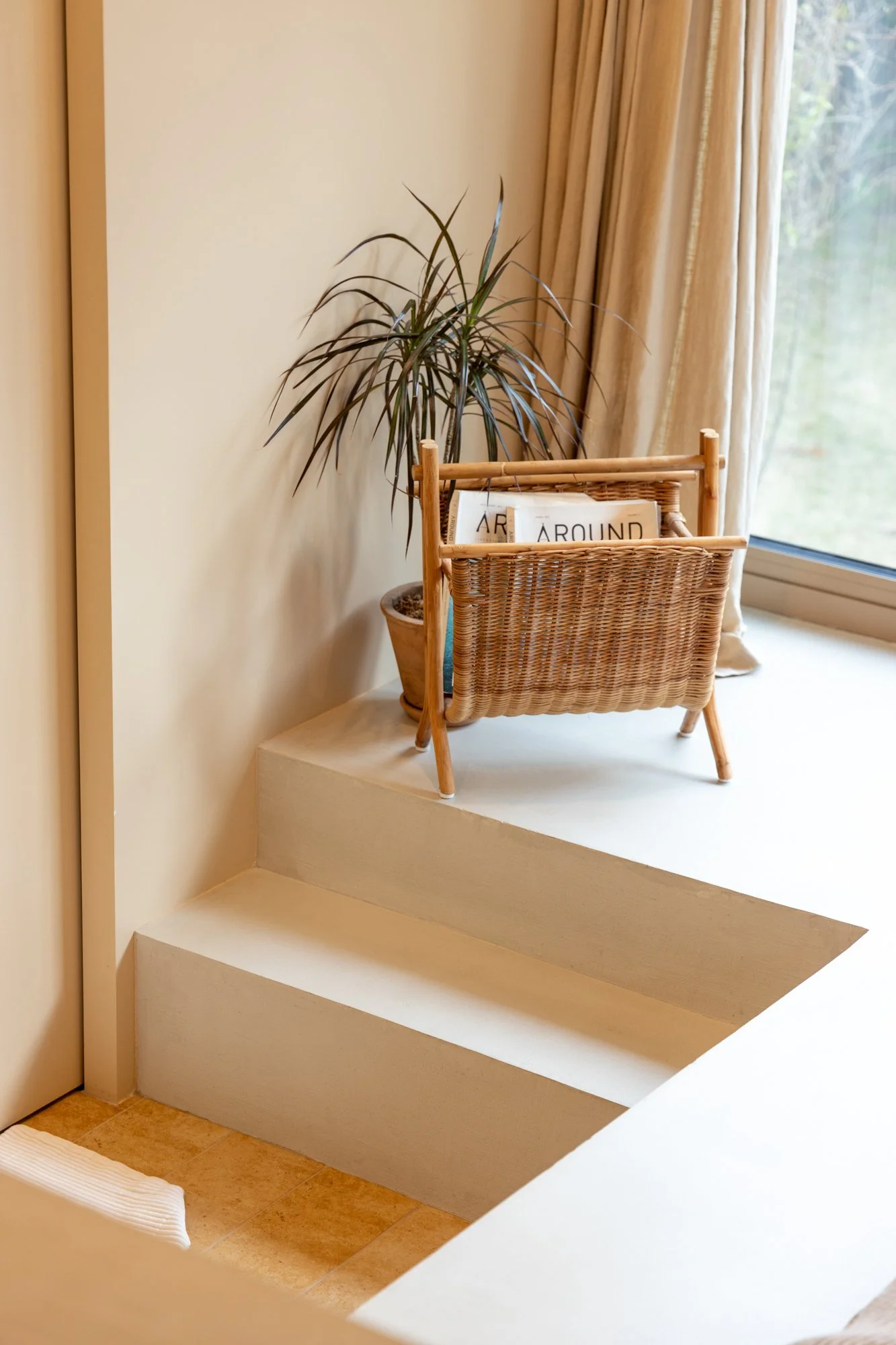 A wicker basket on a white windowsill next to a tall houseplant, with a pot of soil and magazines inside the basket. A window with beige curtains is in the background.