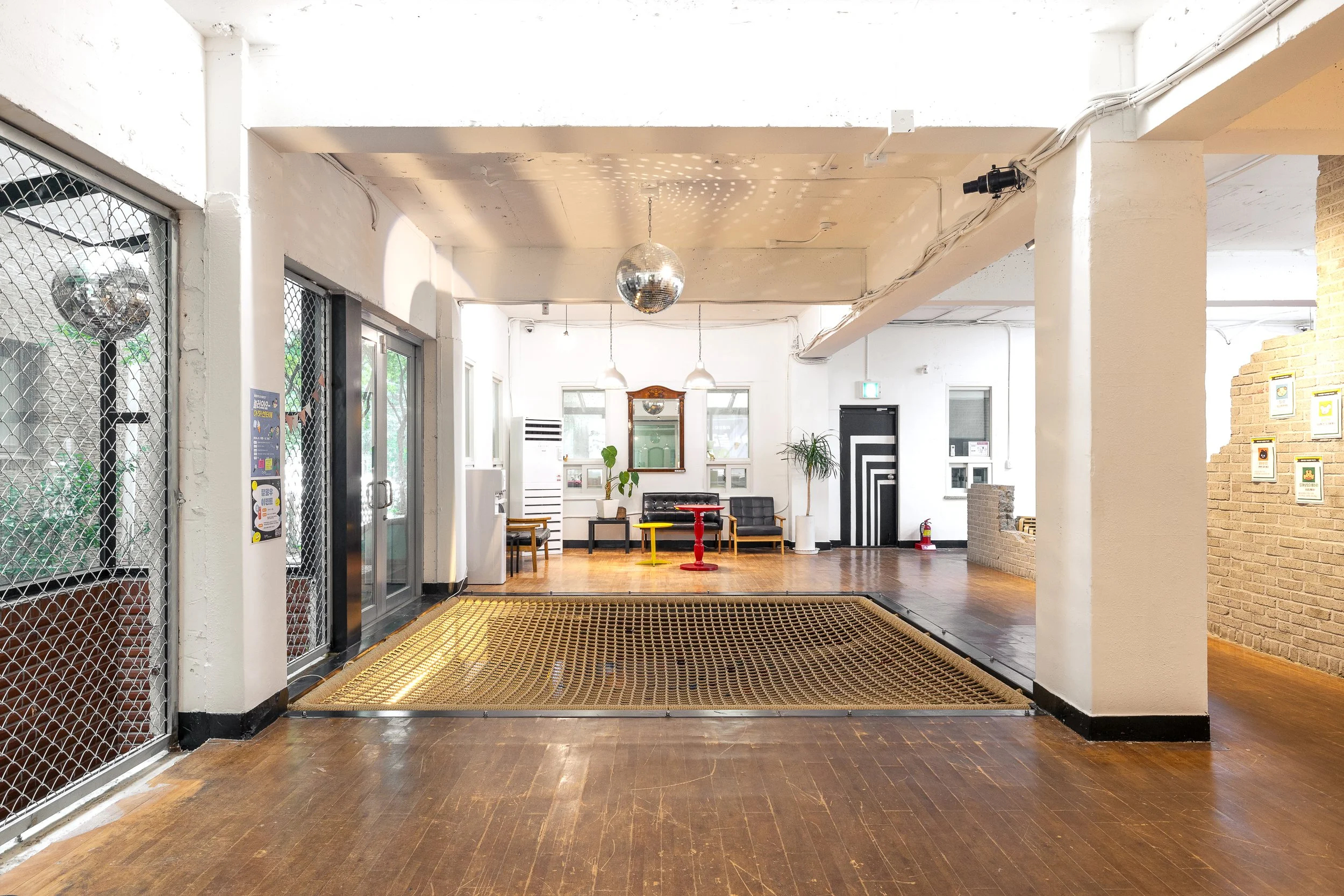 Interior of a modern space with wooden floors, white walls, a black door with a geometric pattern, a small seating area with chairs, a mirror, and a red table, decorated with plants and framed pictures.