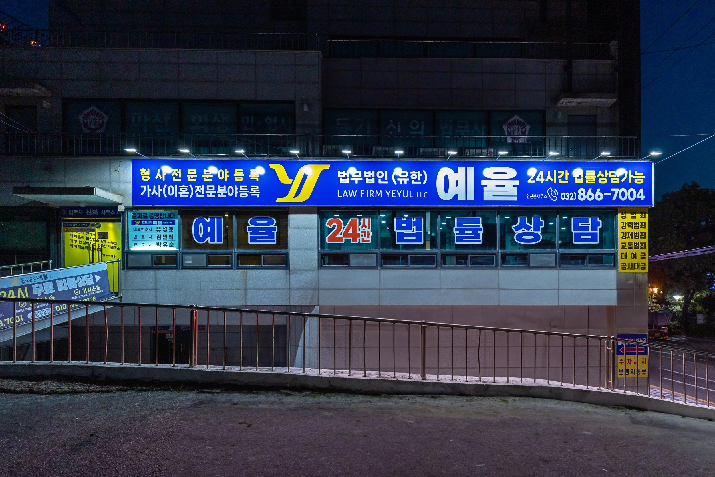 Night view of a building with neon signs in Korean and English advertising a law firm, LAW FIRM YEYULL LLC, available 24 hours with a contact number. The signs are illuminated, and the building has a staircase on the left side. Streetlights and some 