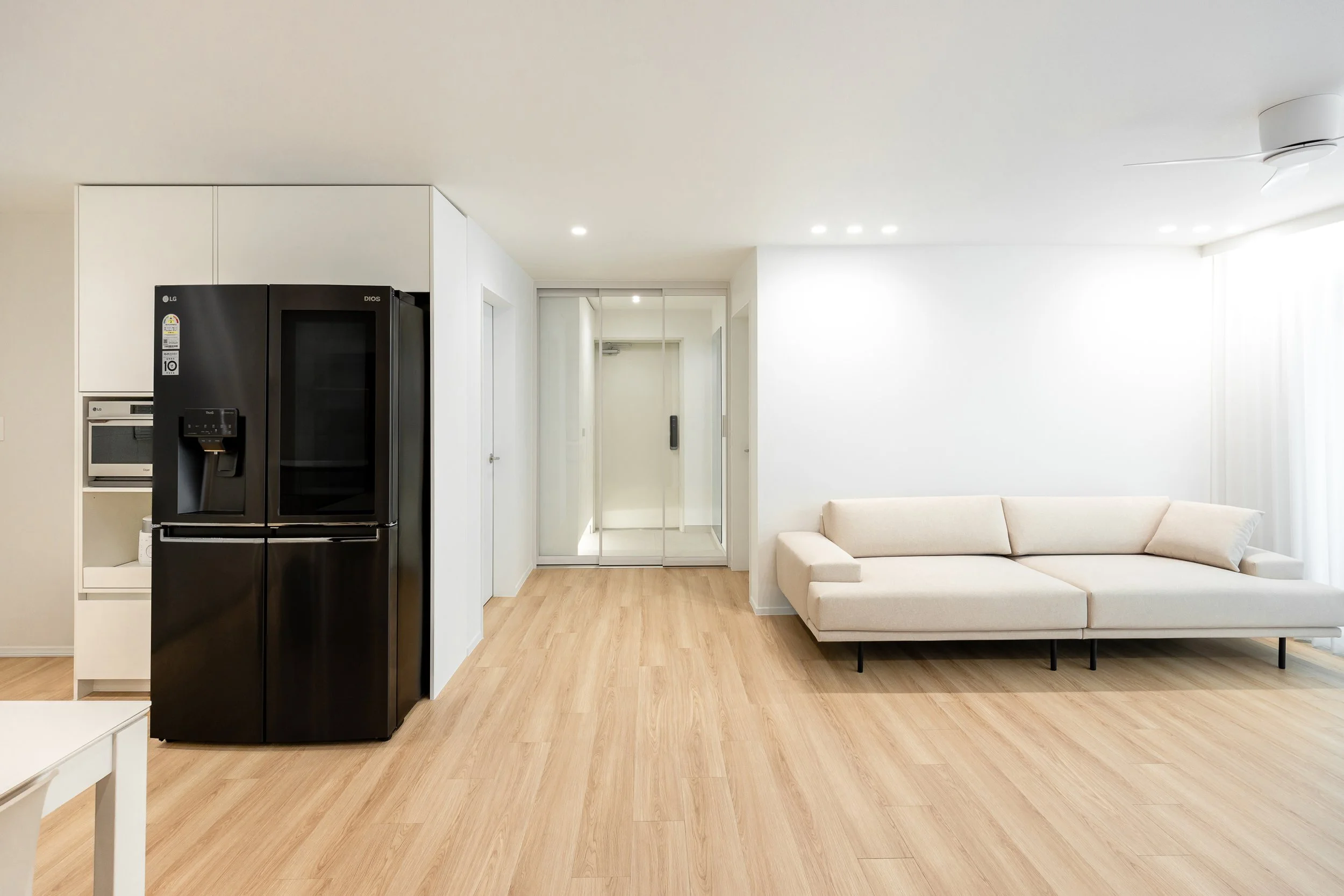 Living room with a white sofa, black refrigerator, microwave, and oven, light wooden flooring, white walls, and glass door to hallway.
