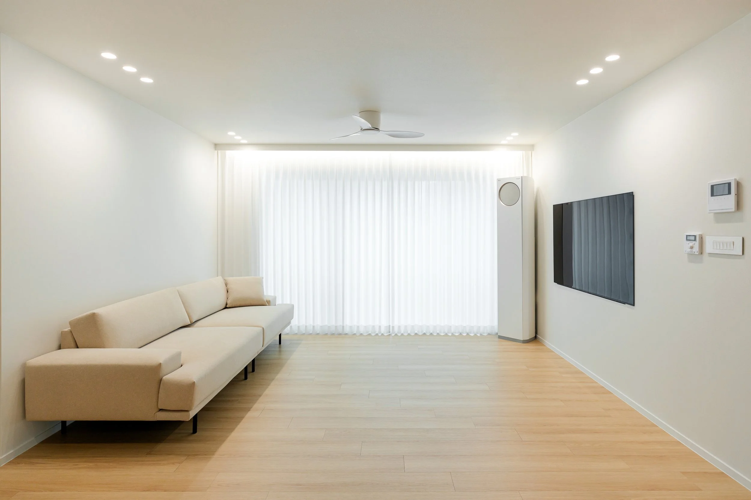 Minimalist living room with beige sofa, large window with white curtains, mounted flat-screen TV, white speaker, and light wood flooring.