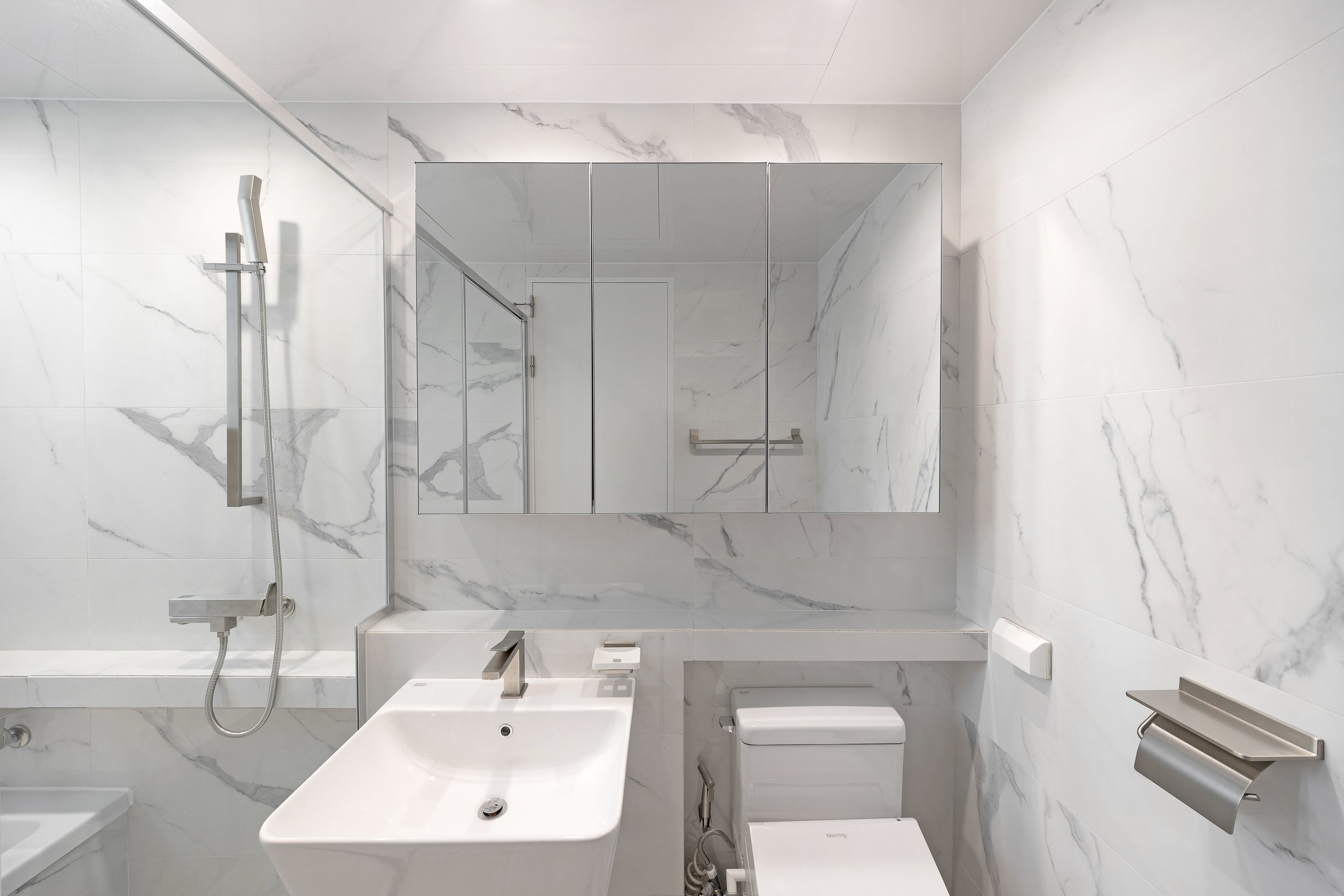 Modern bathroom with marble walls, a mirror cabinet, a sink, a toilet, and a shower area with a sliding glass door.