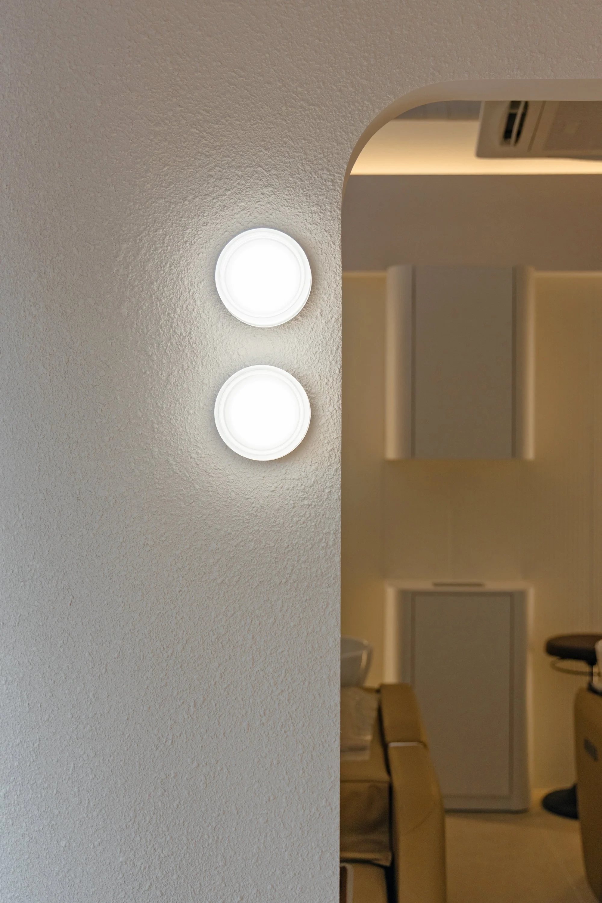 Close-up of two wall-mounted LED lights on a textured white wall in a living space, with part of an interior room visible in the background.