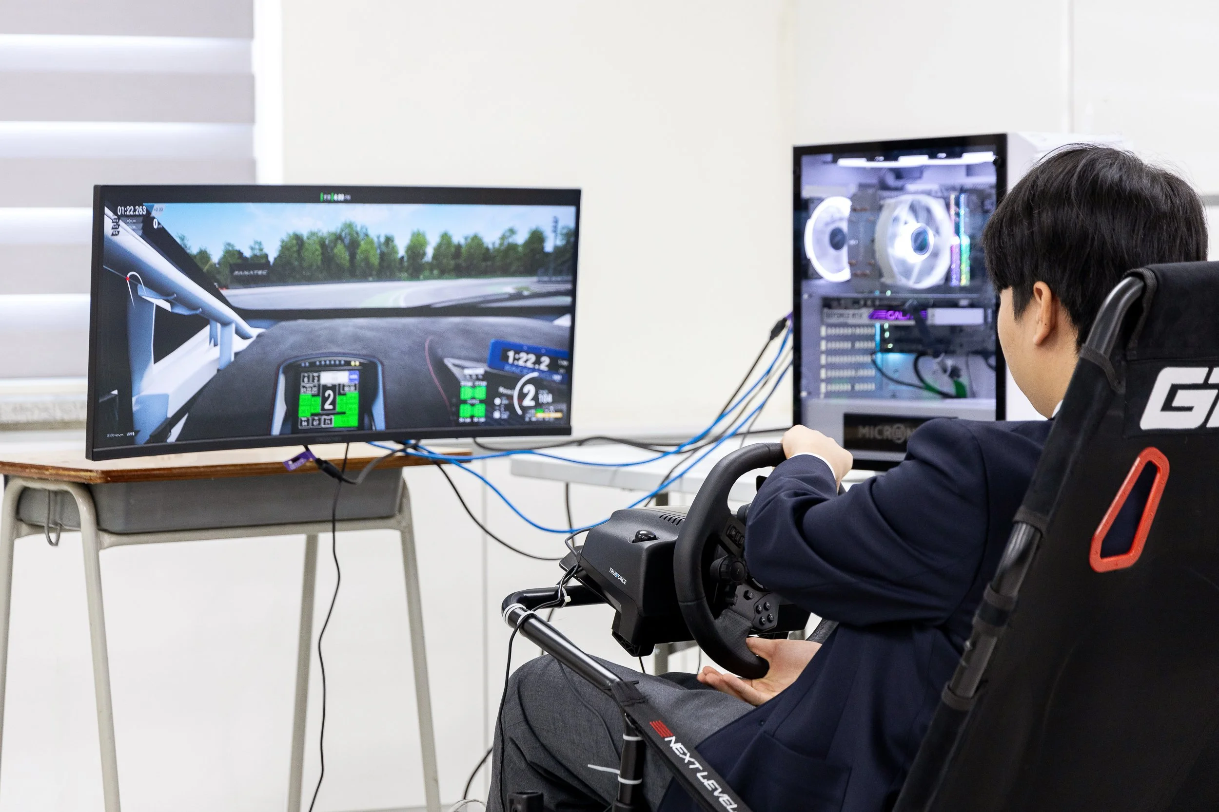 Person in a suit sitting in a racing simulator, holding a steering wheel connected to a computer, with a racing game displayed on a monitor in front of them.