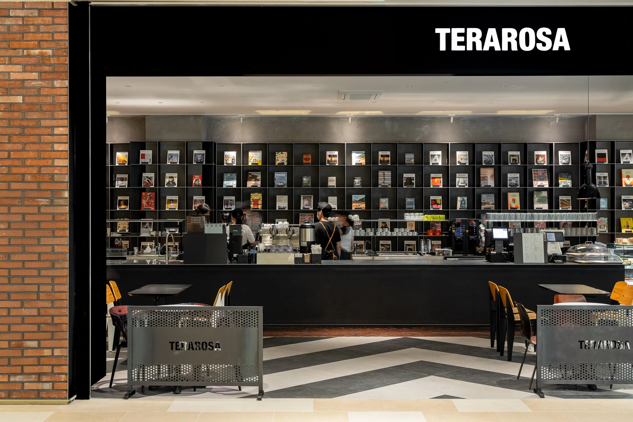Interior view of Terarosa cafe with a black counter, several employees working, and wall shelves filled with magazines or books, in a modern shopping mall.