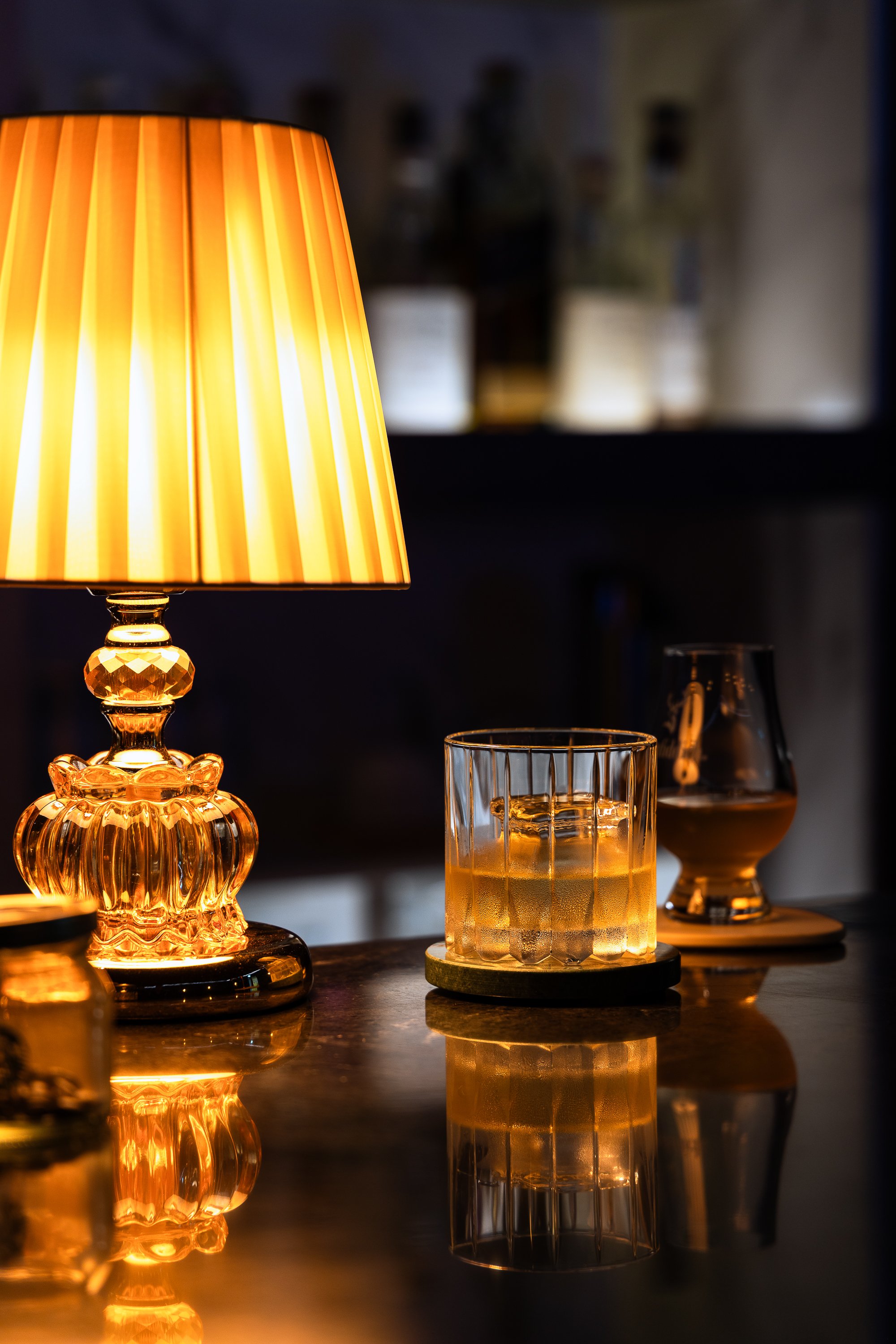 A lit table lamp with a yellow and white striped shade, creating a warm glow. Next to it, there is a glass of whiskey on a coaster, and other glasses containing dark liquids on a polished surface with reflections.