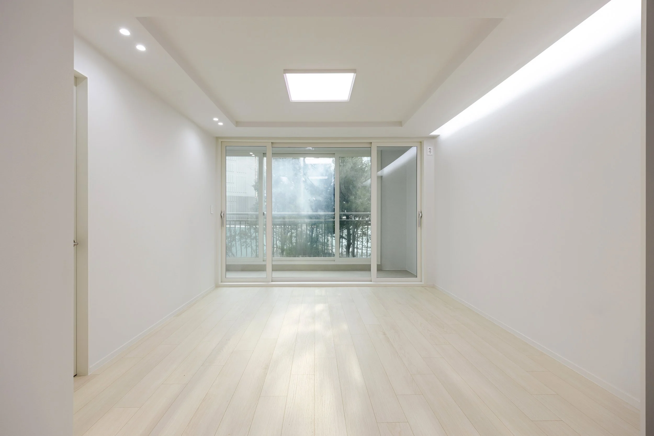 Empty room with white walls, light-colored flooring, large sliding glass door, balcony, and ceiling lights.