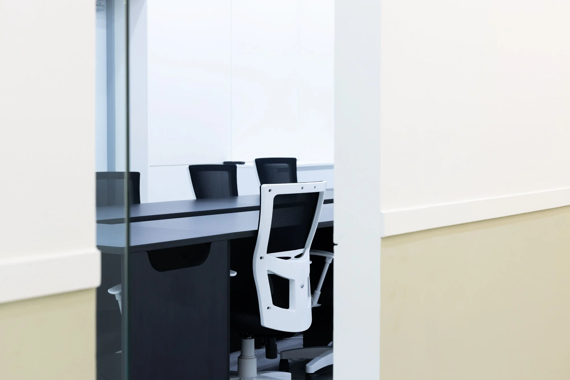 Empty office conference room with black chairs and a white ergonomic chair at a long table, viewed through a doorway.