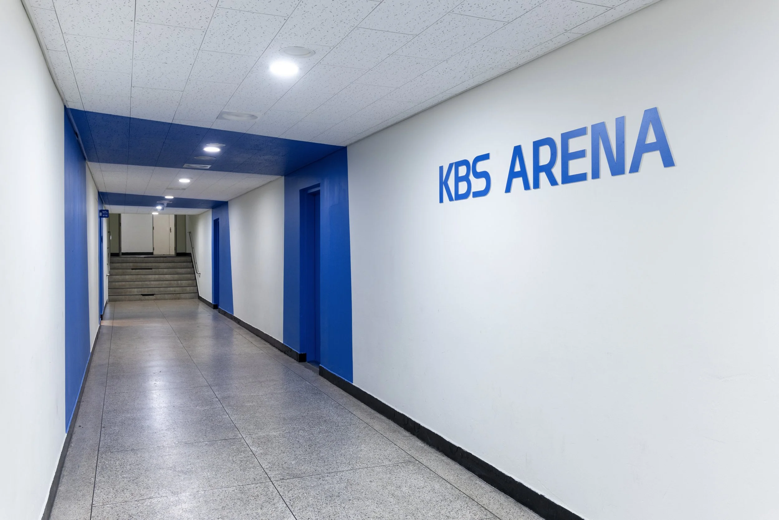 A hallway with white walls and blue accents, leading to stairs, with a sign reading 'KBS ARENA' on the right wall.