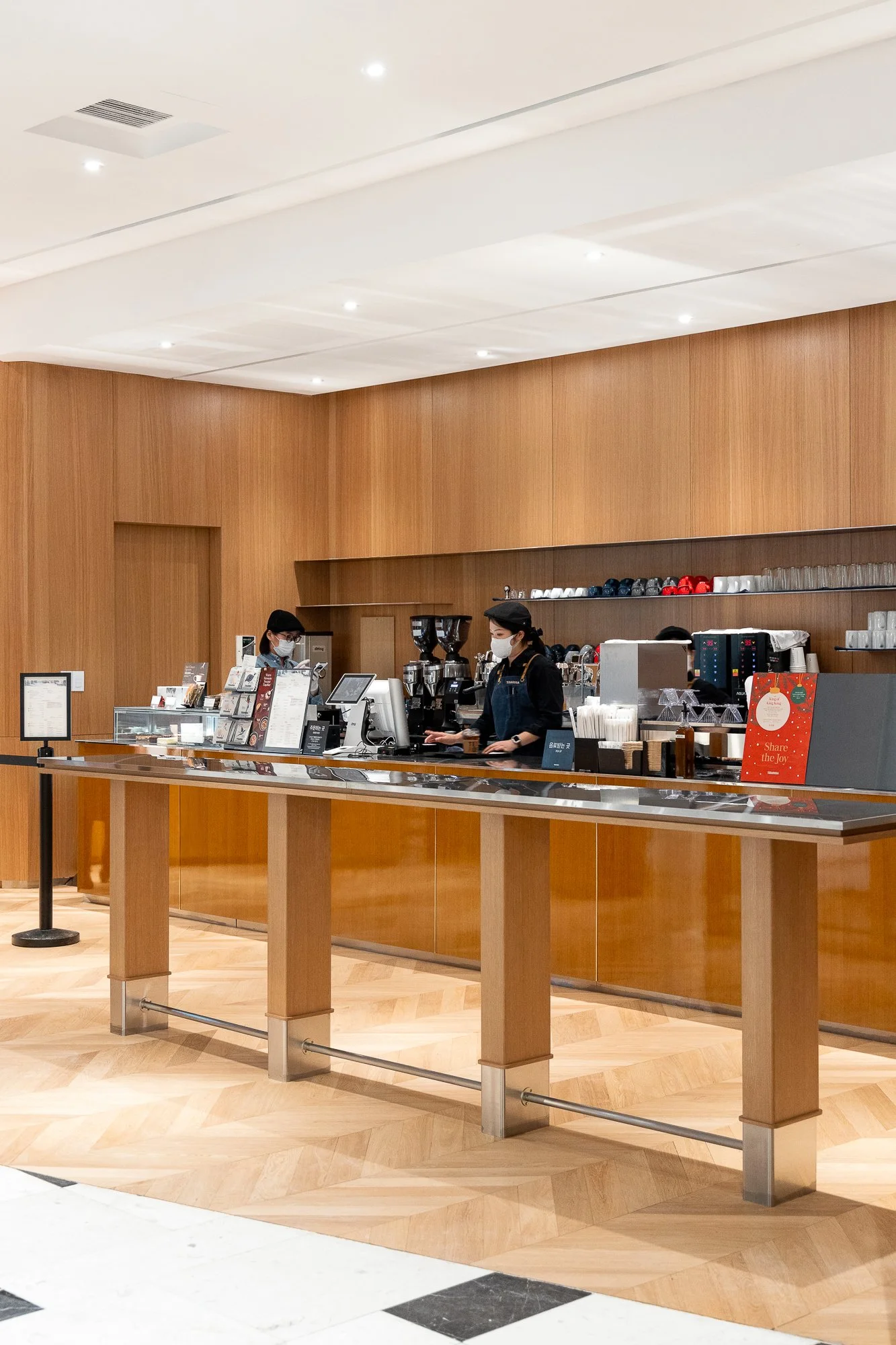 A modern coffee shop counter with two employees wearing masks, black caps, and aprons, preparing drinks behind the wooden counter with coffee machines and cups, inside a well-lit, wooden-paneled cafe.