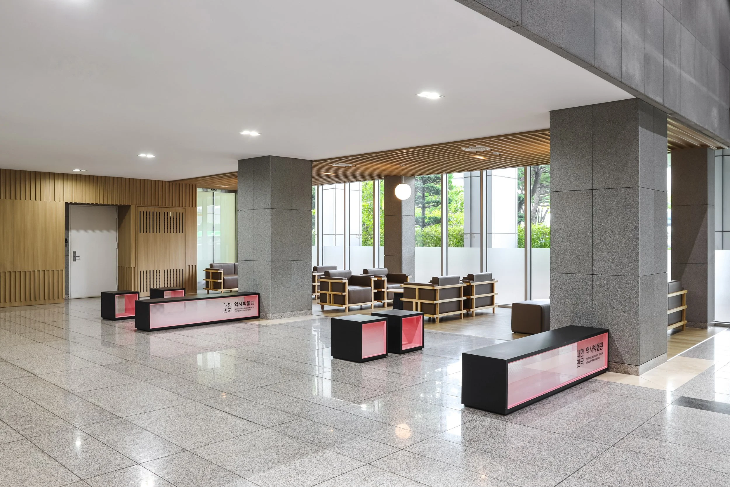 A modern indoor lobby area with large windows, gray columns, wooden ceiling accents, seating with brown chairs and gray cushions, and black benches with pink lights.