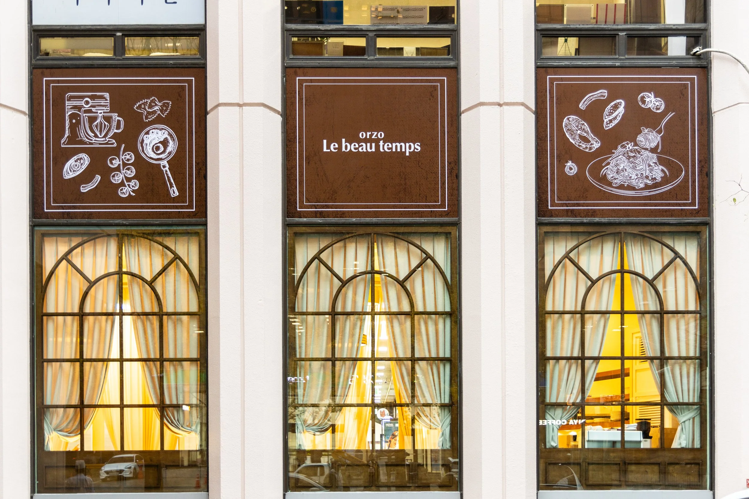 The exterior of a restaurant with three large windows, each adorned with yellow curtains and view inside, illuminated with warm lighting. Above the windows are three square panels with brown backgrounds and white line drawings of kitchen utensils and