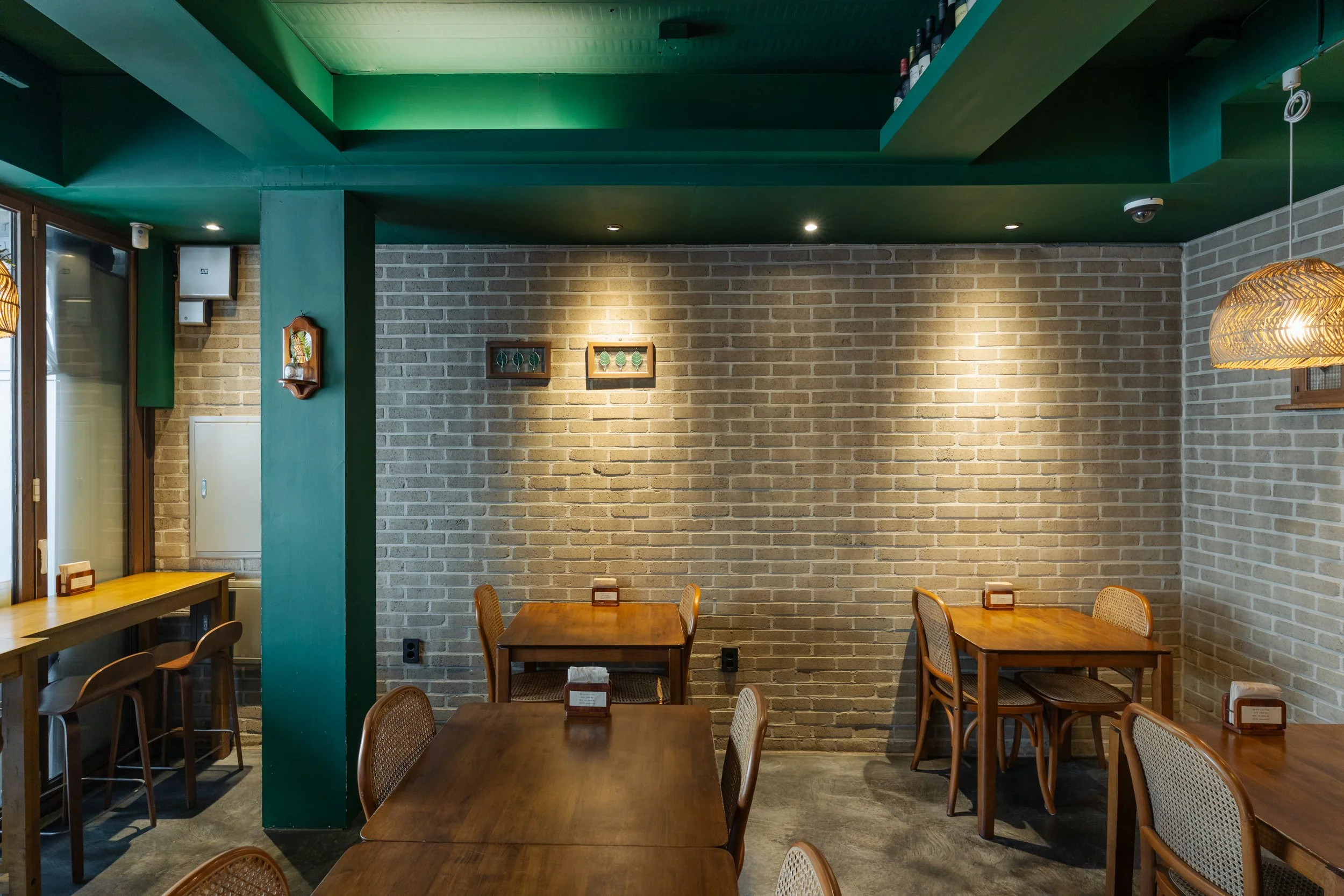 Interior of a small restaurant with wooden tables and chairs, brick walls, green ceiling accents, hanging wicker lamps, and framed artwork.