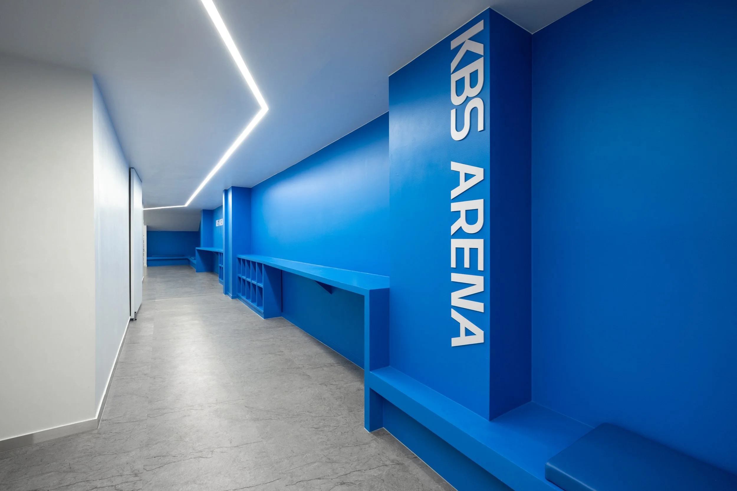 Modern indoor hallway with a blue accent wall featuring white text 'KIBS ARENA' and a bench below, illuminated by sleek fluorescent lighting.