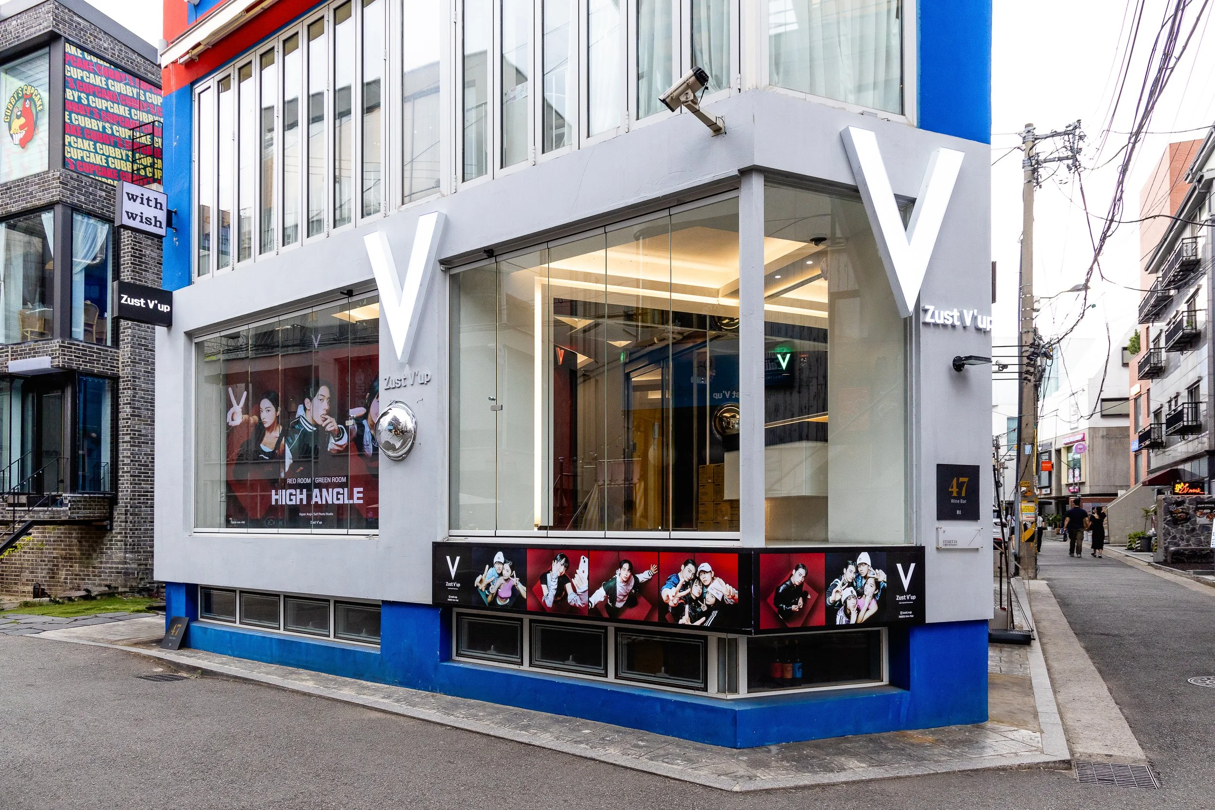 Modern two-story building with large glass windows, displaying promotional posters of urban youth, and illuminated white letter V signs on the exterior. The building is located on a street corner with neighboring buildings and power lines visible.