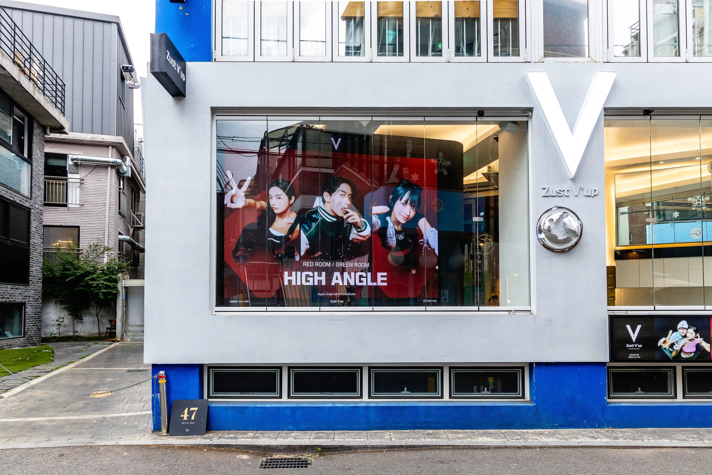 Exterior of a modern building with large glass windows displaying a poster of three young people, advertising a photo studio called High Angle, with a sign reading 47 Wine Bar in front.