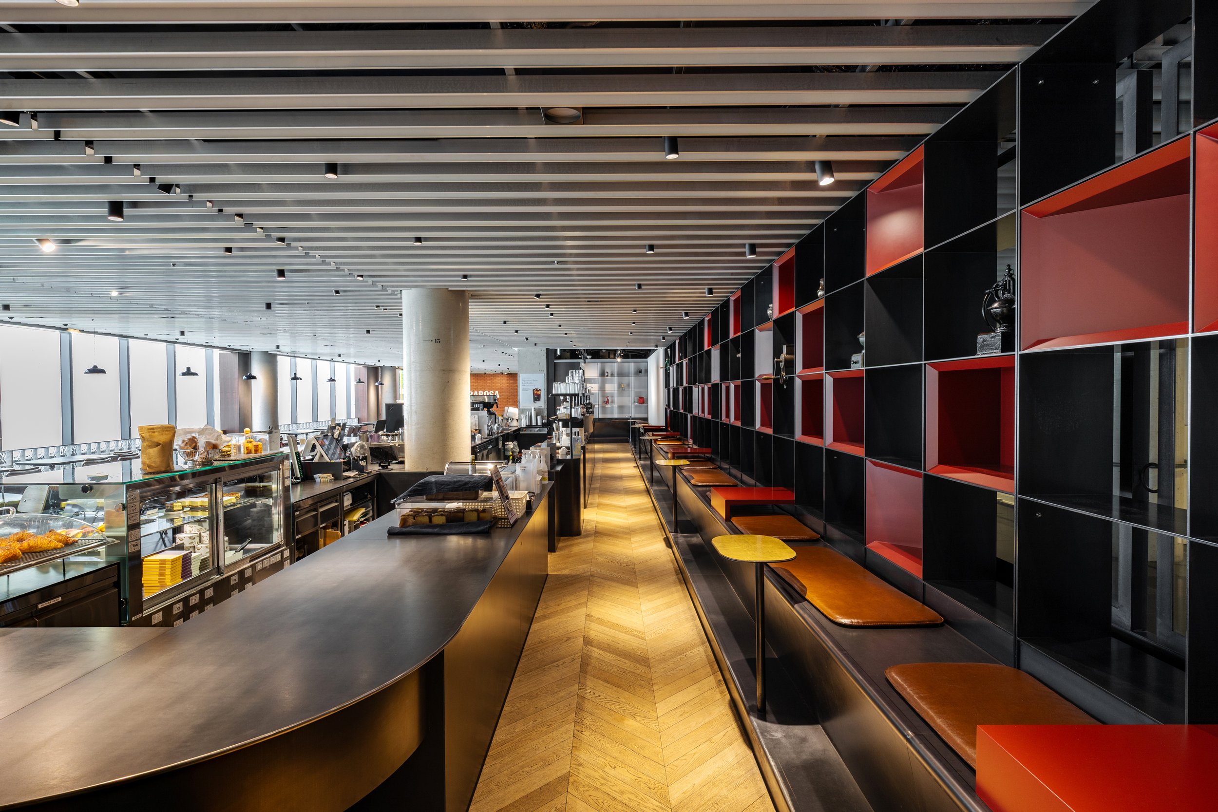 Interior of a modern restaurant or cafe with a long counter and a seating area with wall shelving that has decorative items. Large windows provide natural light.