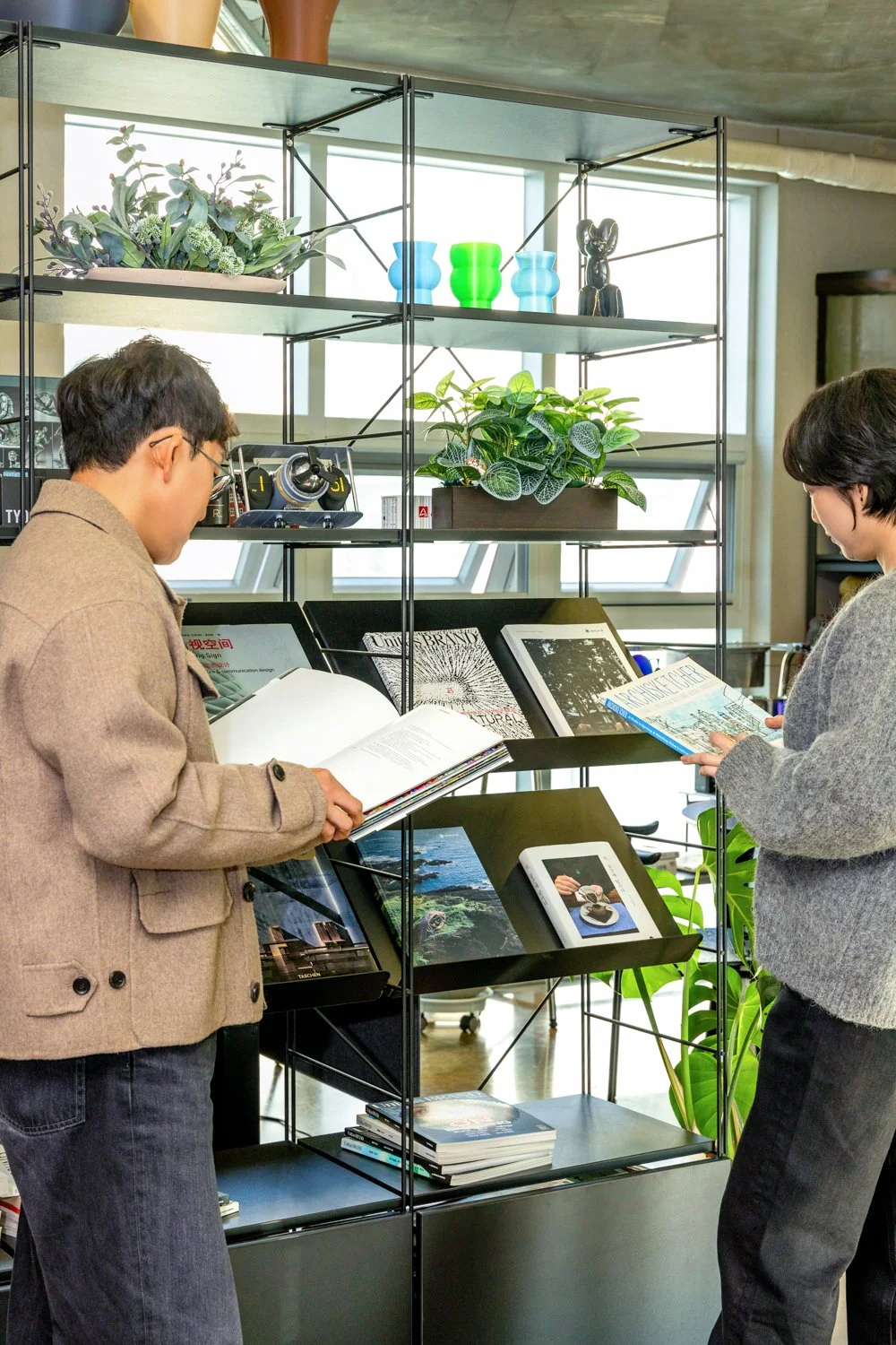 Two women browsing books and magazines in a modern indoor space with shelves and plants.