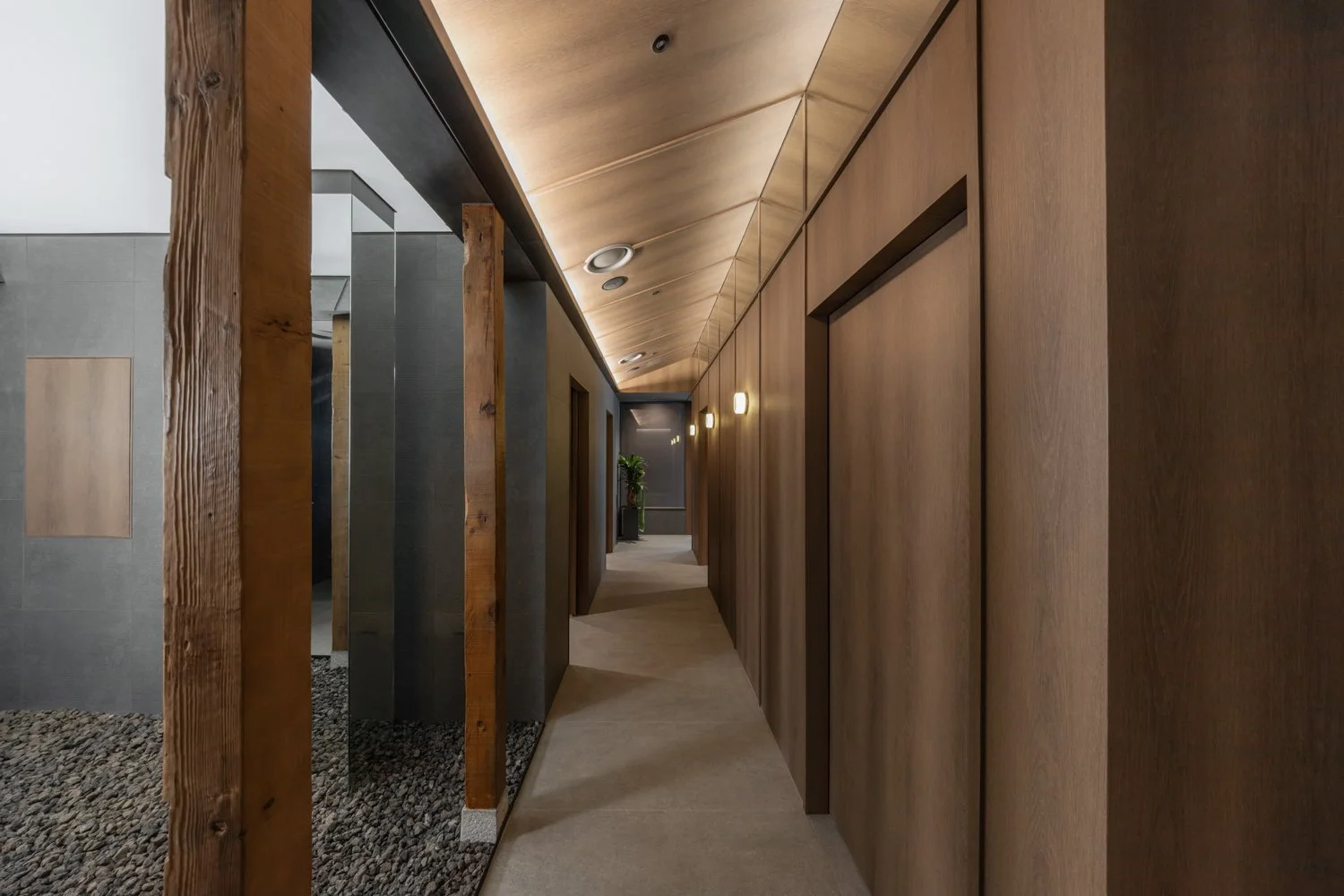 A modern hallway with wooden panel walls and a sloped wooden ceiling illuminated by built-in lights. On the left, a partial wall with black tiles and wooden beams, with gray stone flooring and small rocks at the base. In the distance, a potted plant is visible.
