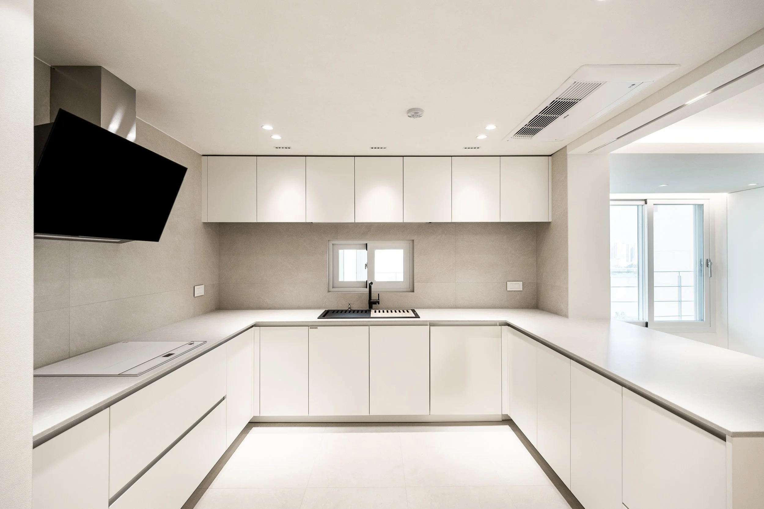 Modern minimalist kitchen with white cabinets, light gray countertops, small window above the sink, black faucet, and a wall-mounted TV in the corner.