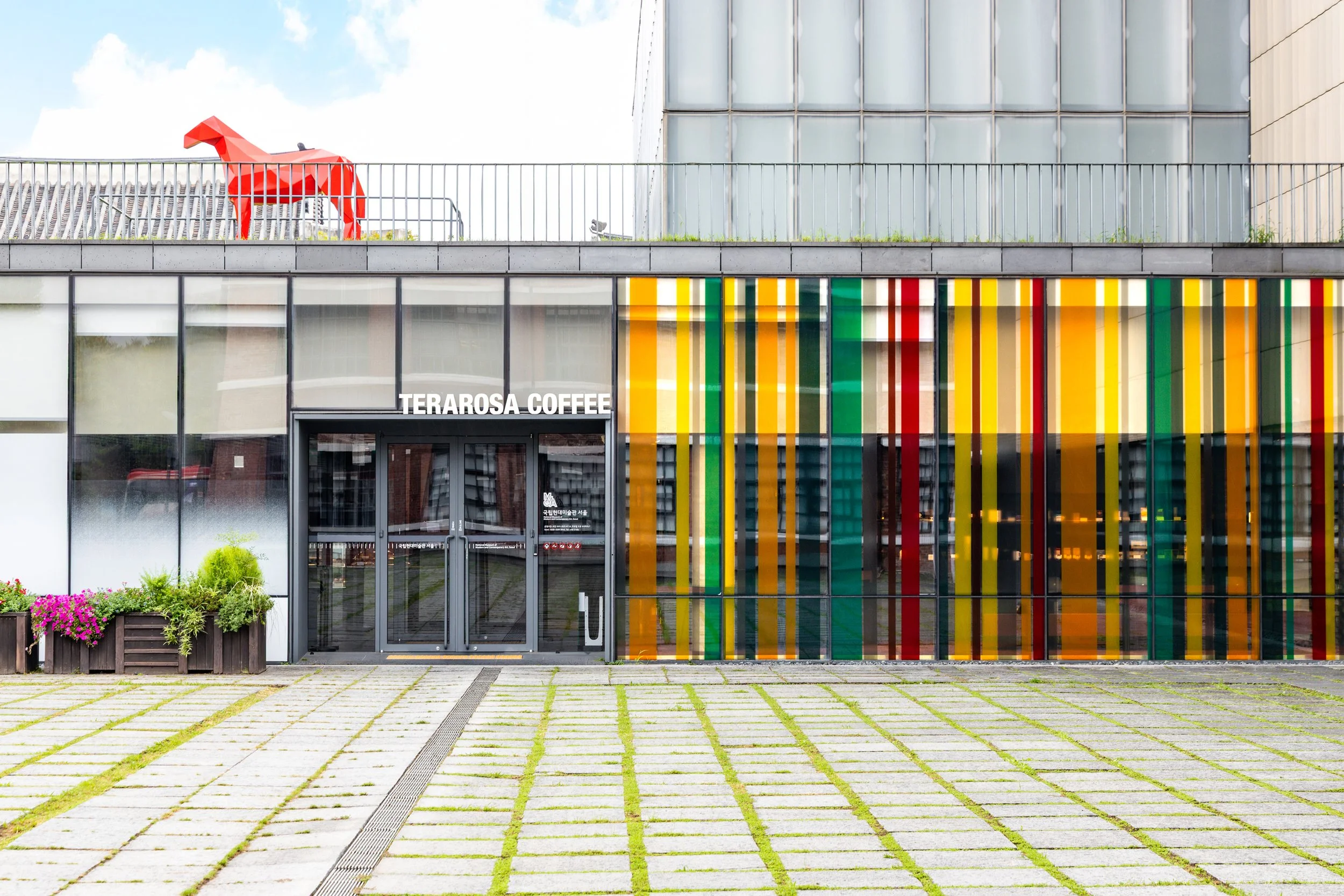 Modern building with colorful vertical striped glass panels on right side, a clear glass door entrance labeled 'TERAROSA COFFEE', with flower planters on left, and a rooftop sculpture of a red horse on top of the building.