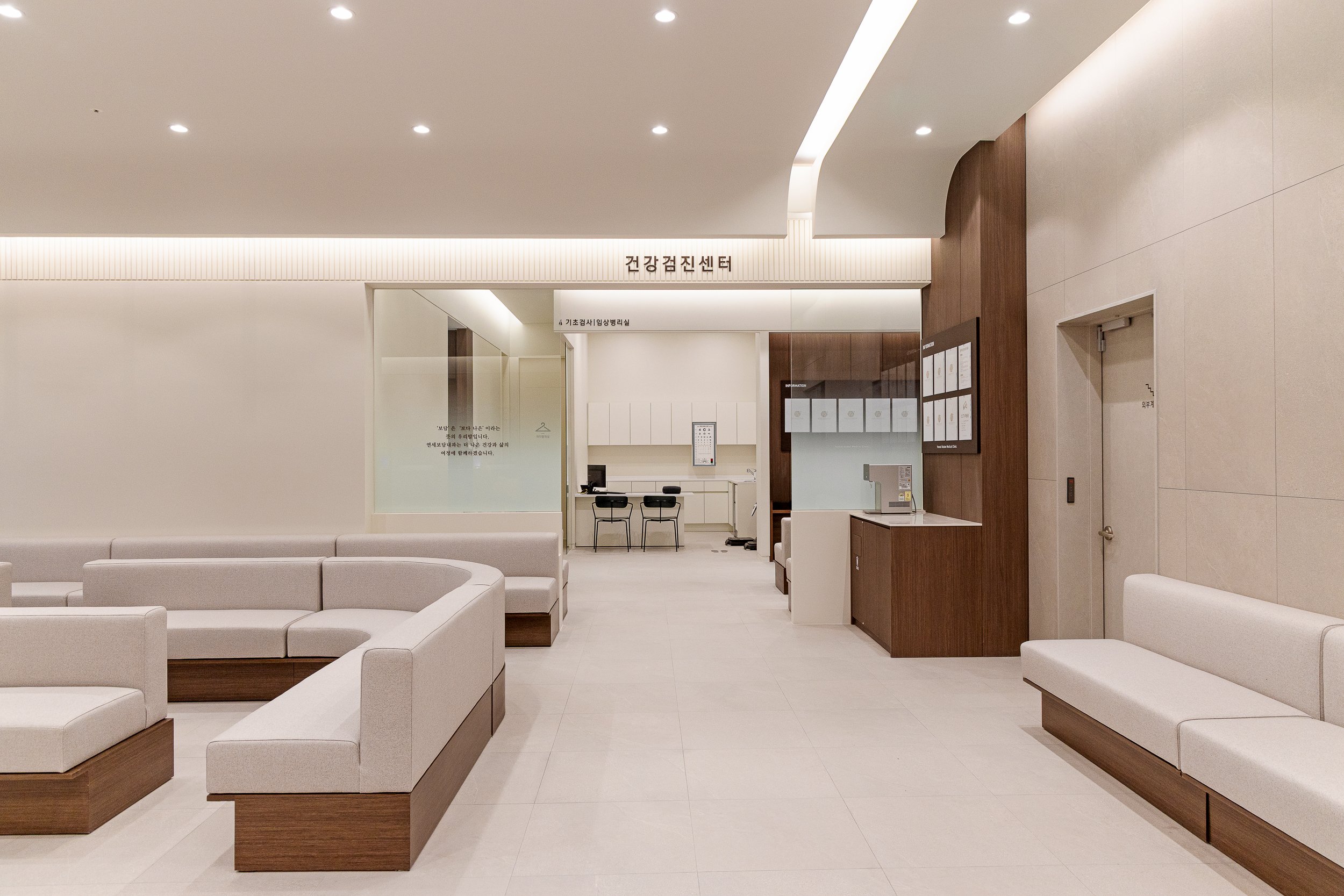 Empty hospital waiting area with white and beige seating, wooden accents, and a reception counter with chairs and a clipboard on the wall. Signage in Korean above the counter indicates it is a health screening center.