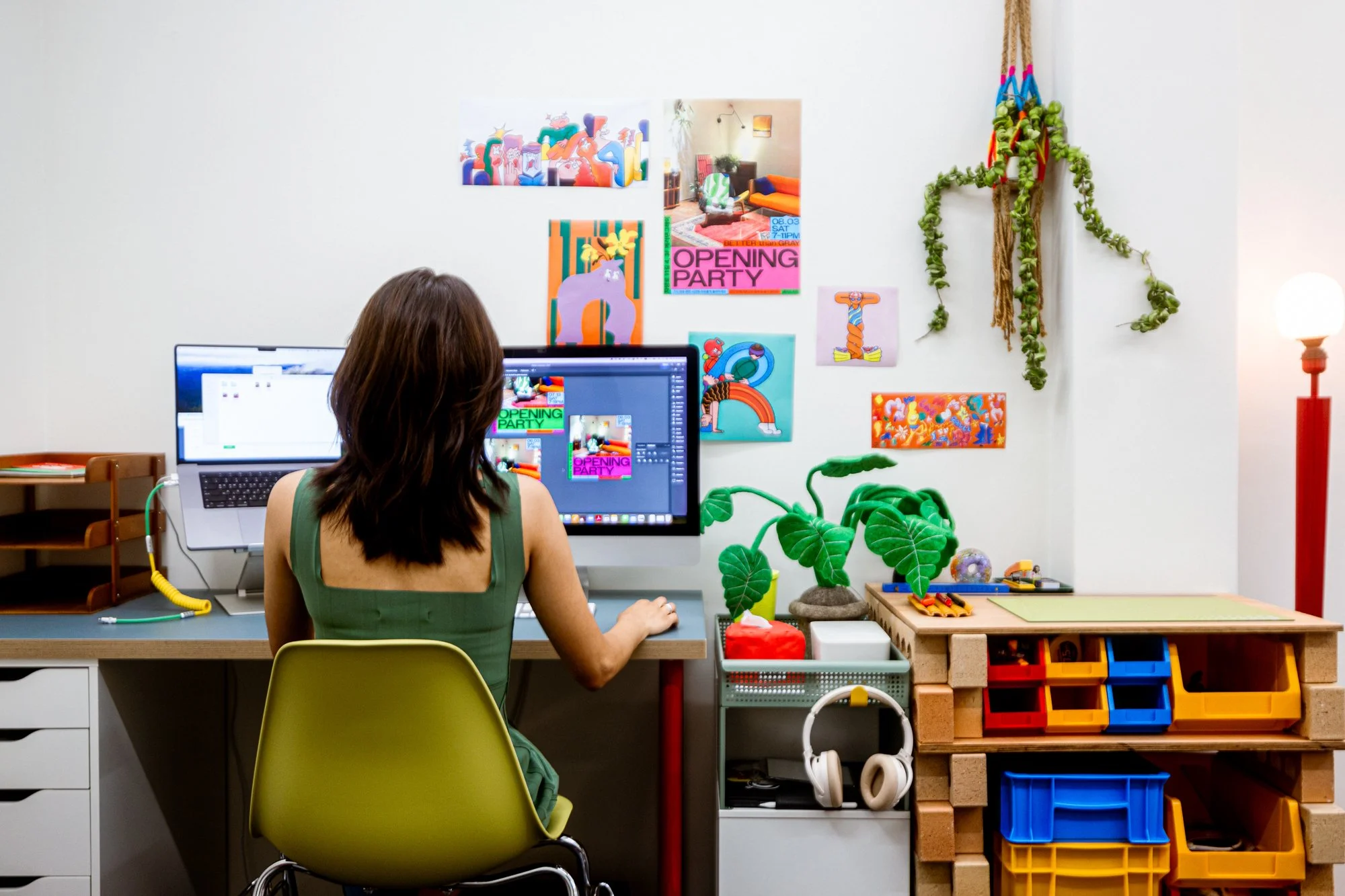 A woman working on a computer at a desk decorated with colorful artwork and plants, with shelves and storage boxes nearby.