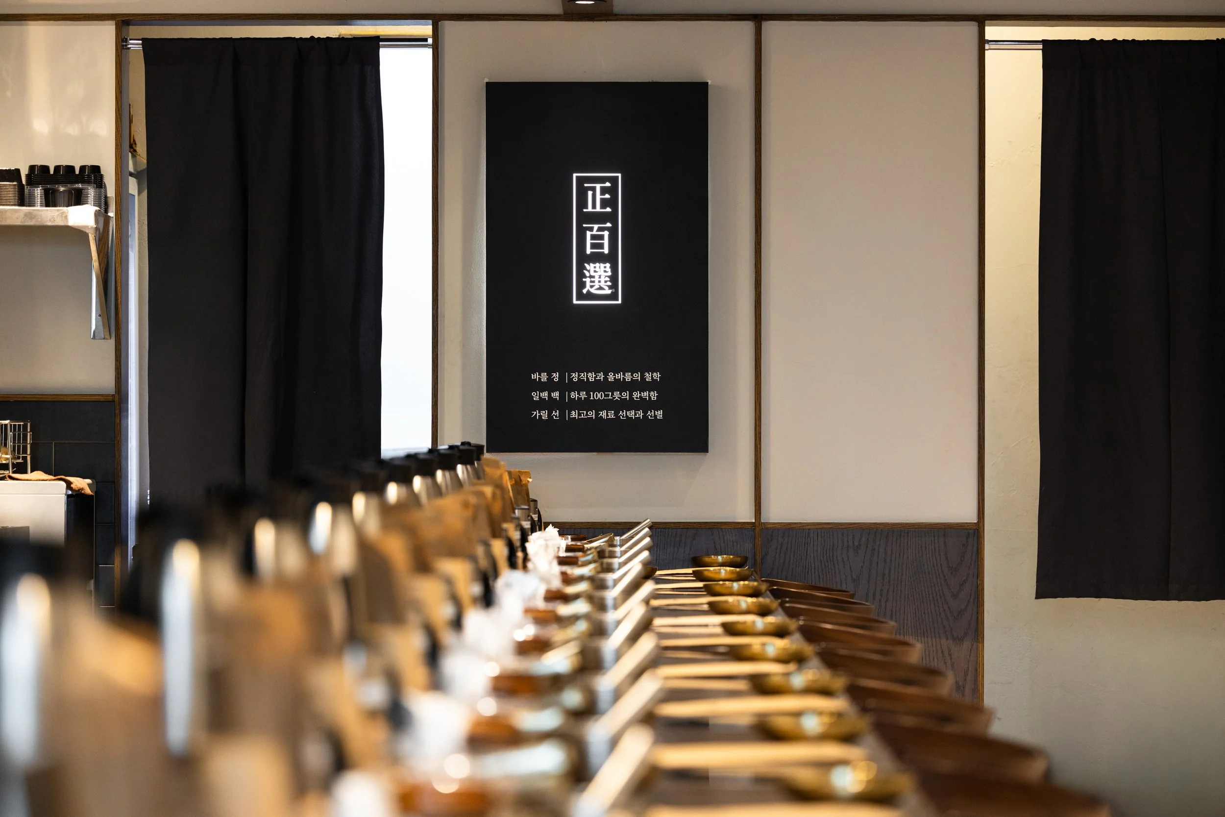 Row of empty traditional Korean wooden bowls and spoons on a table inside a restaurant with black curtains and a black sign with white Asian characters on the wall.
