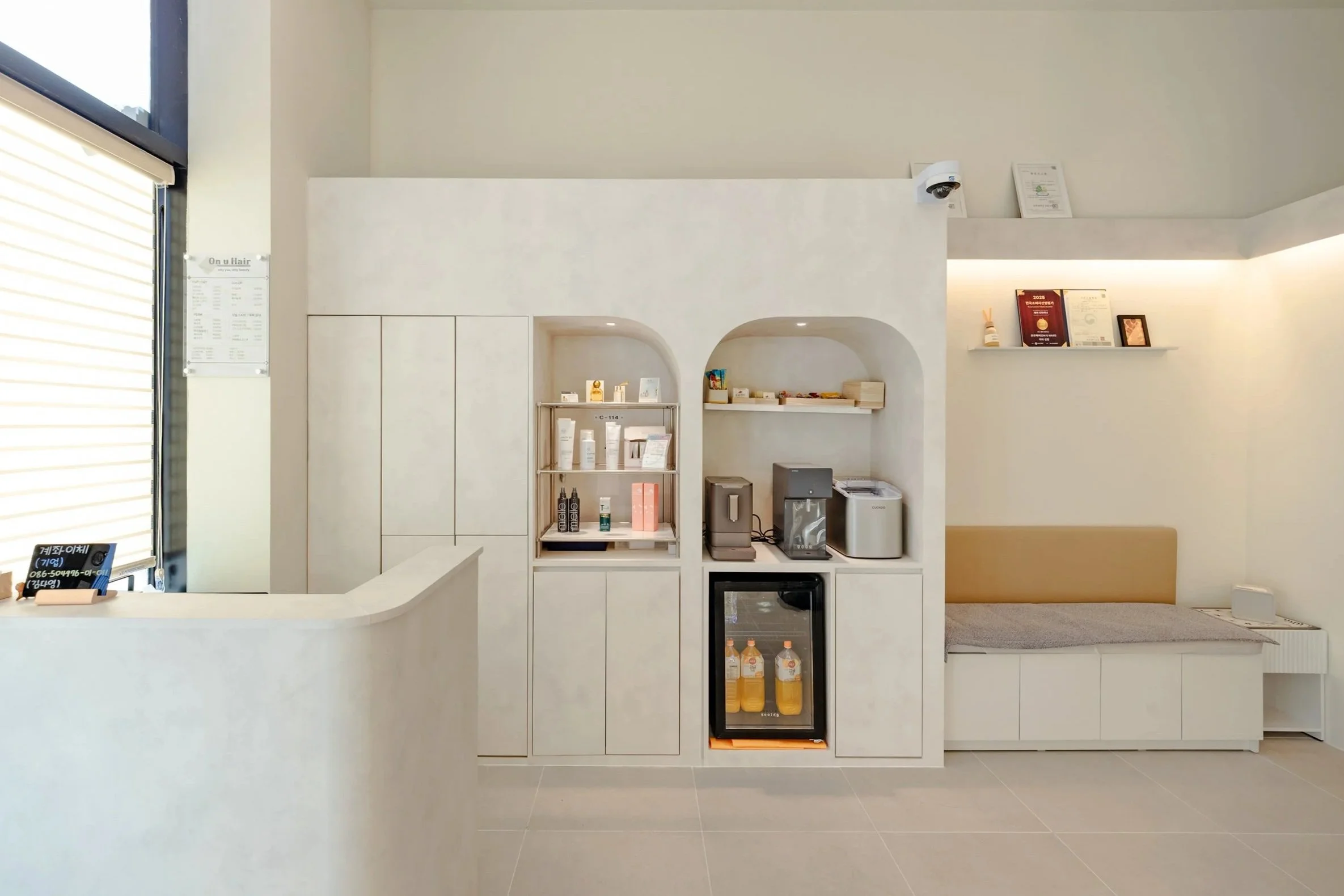 Minimalist reception area with a white counter, a small fridge with drinks, a coffee machine, and a seating nook with a beige cushion. Shelves above hold various items including certificates and decor.