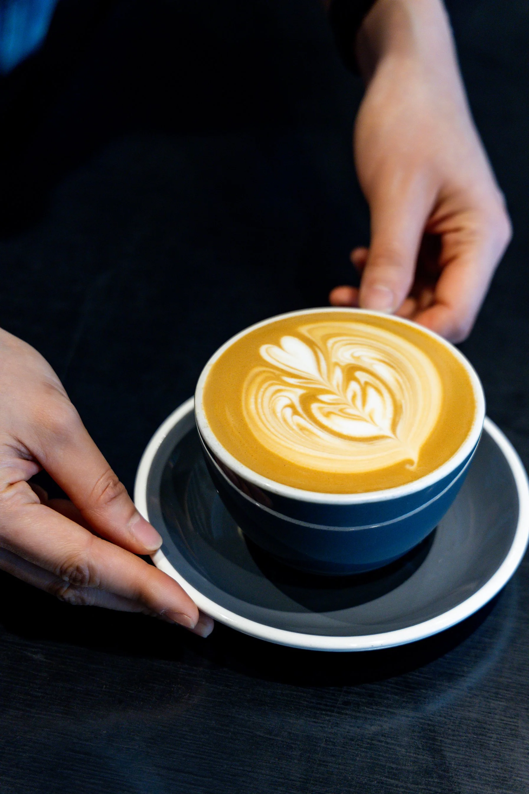 A person holding a cup of coffee with latte art, on a saucer, on a dark table.