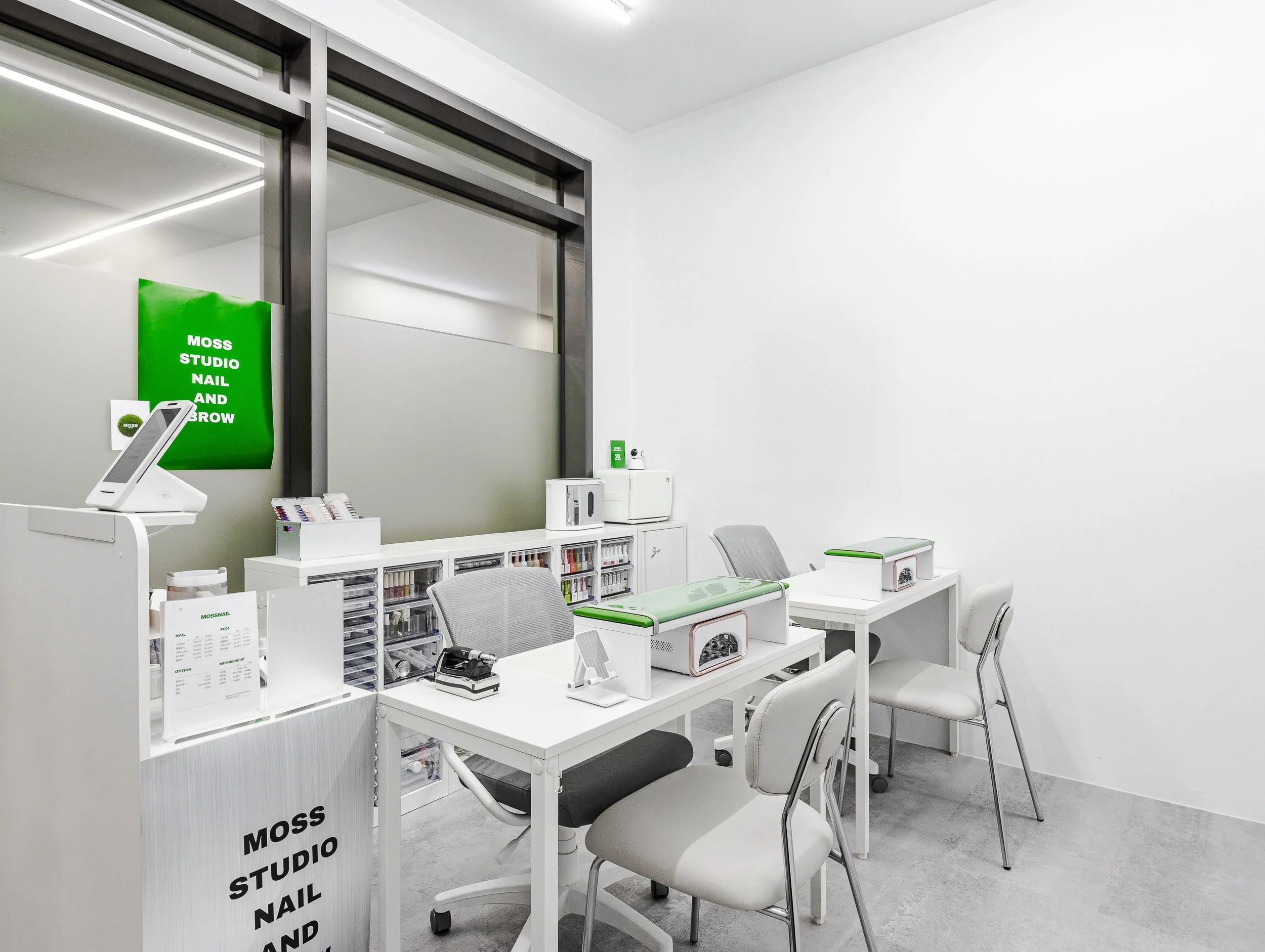 A small, modern nail and eyelash salon with white walls, gray chairs, and manicure tables with green accents. There are storage drawers, a monitor, and salon equipment visible in the space.