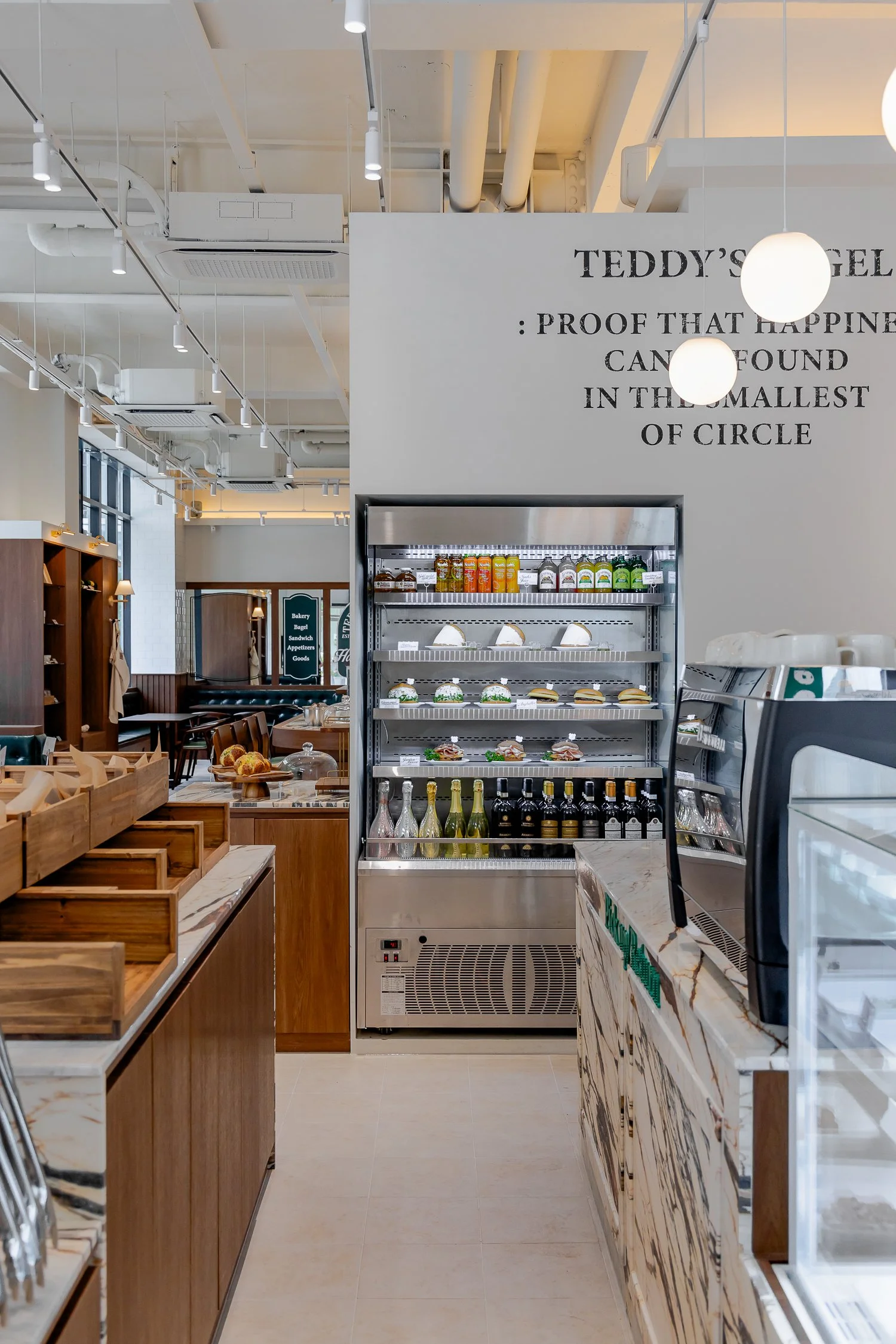 Inside a modern cafe or bakery with a marble counter, wooden shelves, and a refrigerated display with bottled drinks and food items. Text on the wall reads 'Teddy's Eatery: Proof That Happiness Can Be Found in the Smallest of Circles.'