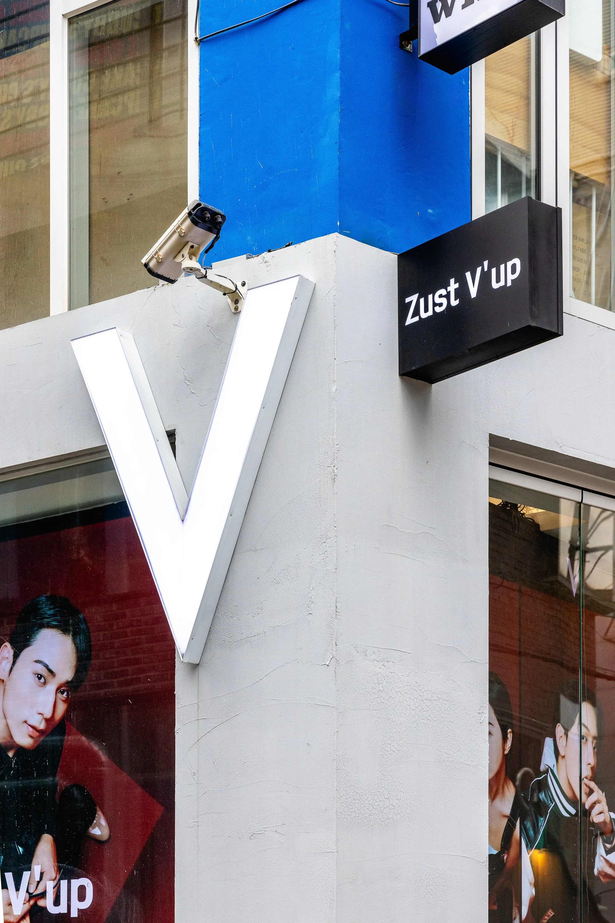 Close-up of a corner of a building with a white wall, a large illuminated white letter 'V', a security camera, and a black sign that reads 'Zust V'up'. There are posters with images of people inside the building.