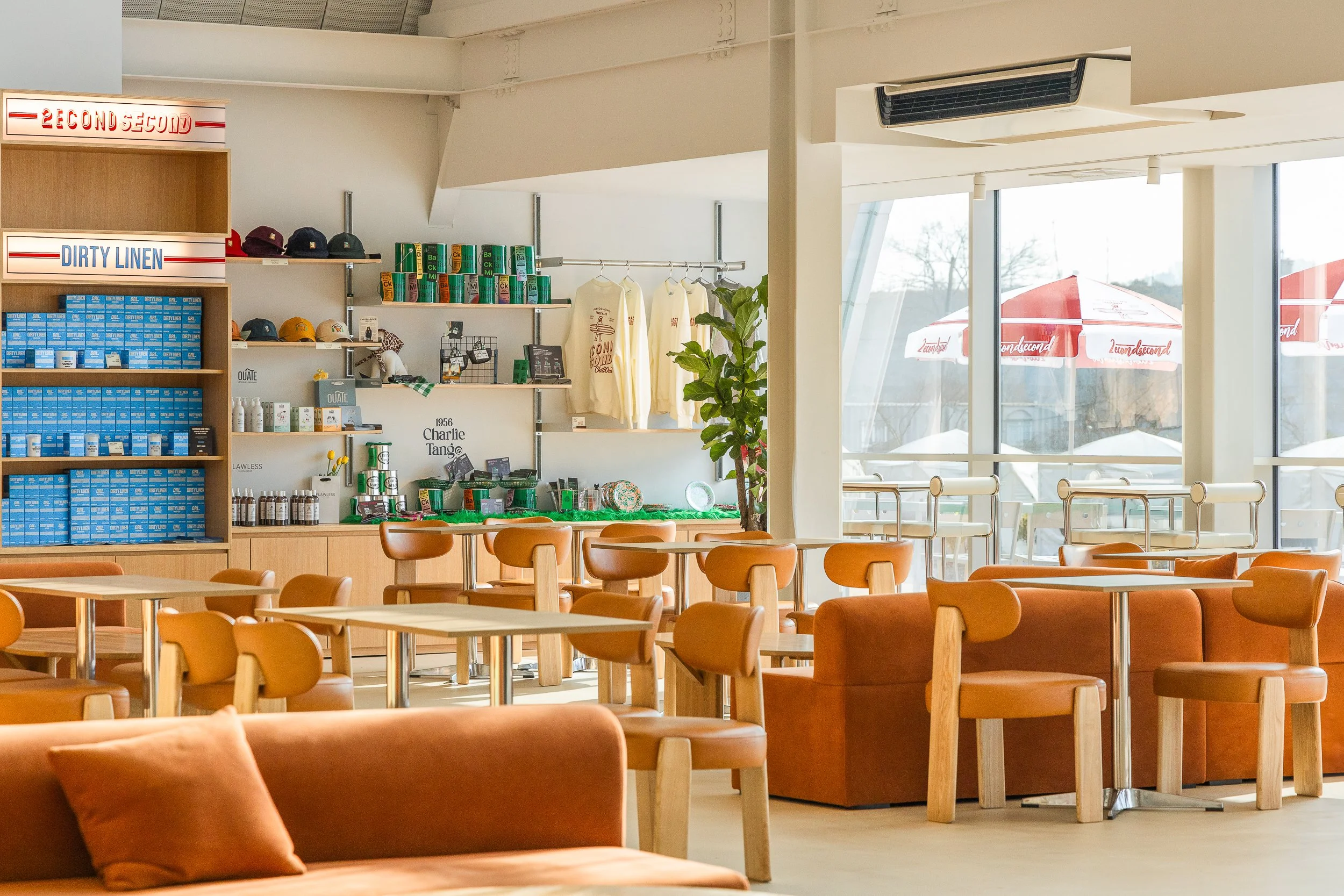 Empty cafe with brown chairs, a sofa with a pillow, large windows with umbrellas outside, and a retail display shelf with caps, shirts, and merchandise.