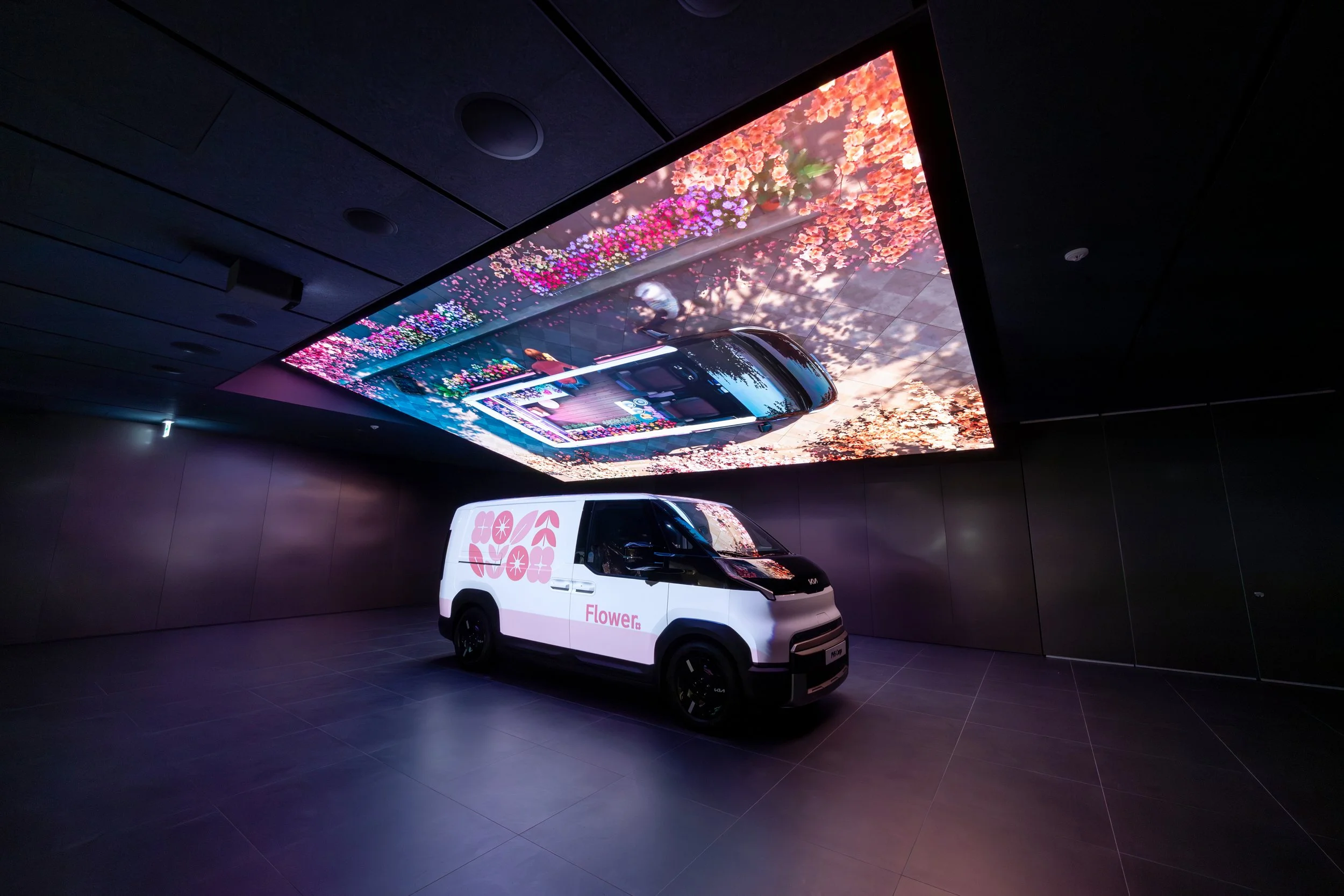 A white delivery van with pink flower graphics and the word AI Flower on the side, placed in a dark room with a large digital screen on the ceiling displaying a colorful flower garden scene. The scene is reflected on the van's windshield.