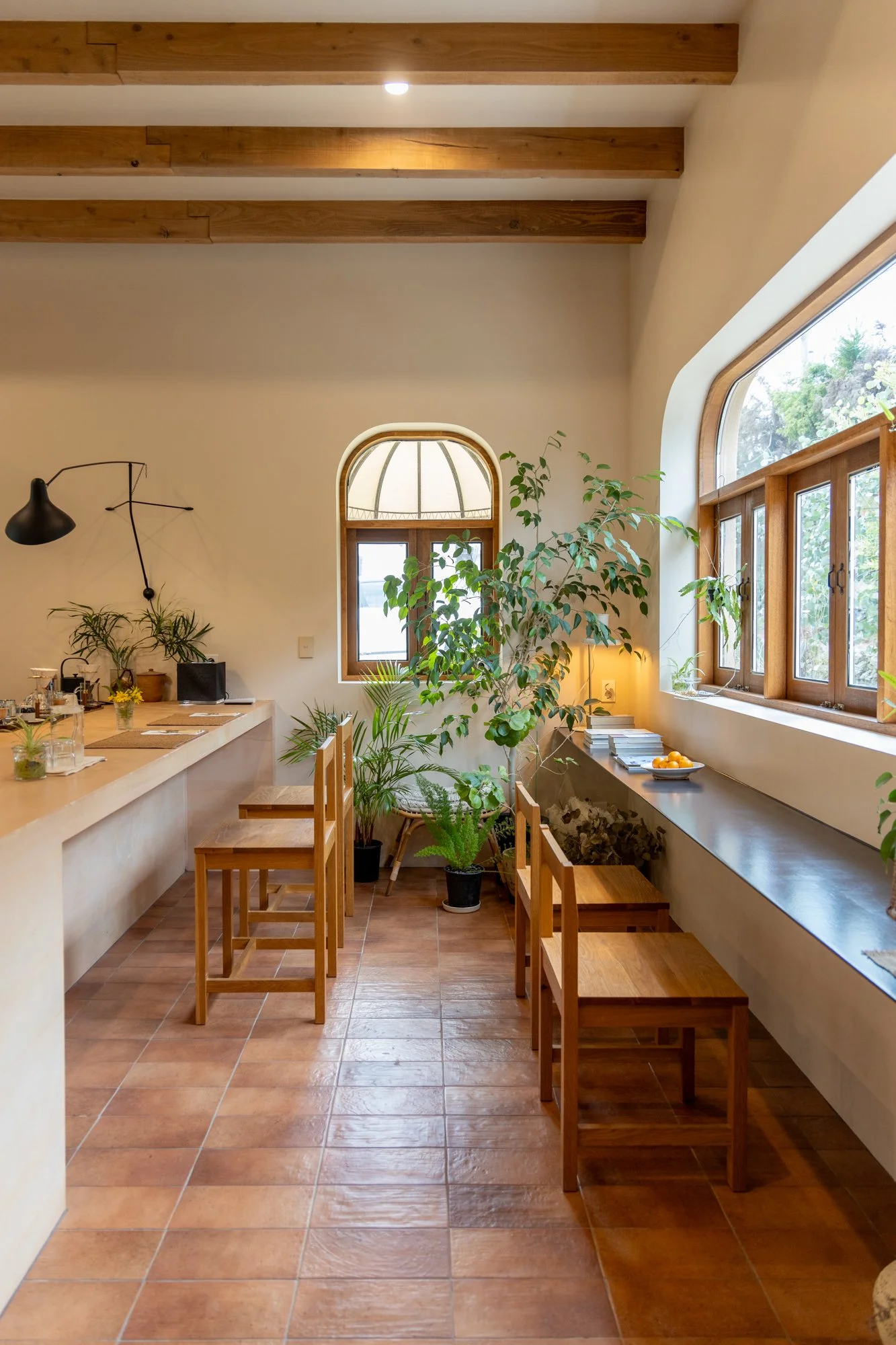 A cozy kitchen corner with terracotta tile flooring, wooden ceiling beams, large windows with wooden frames, a long black countertop, and various potted plants.