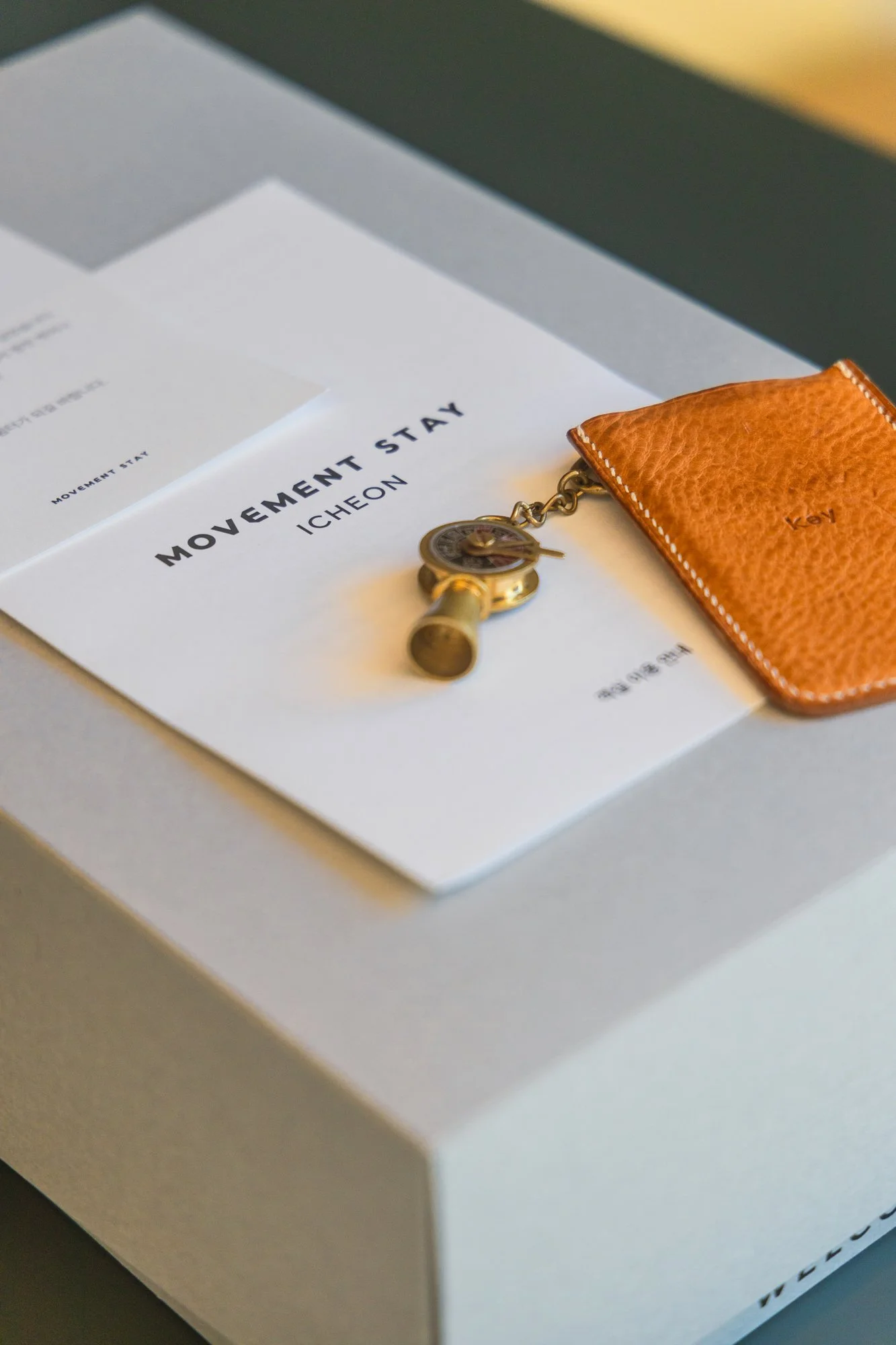 A white box containing a paper titled 'Movement Stay' by Icheon, a gold wristwatch, and a brown leather key holder with the word 'Key' engraved on it.