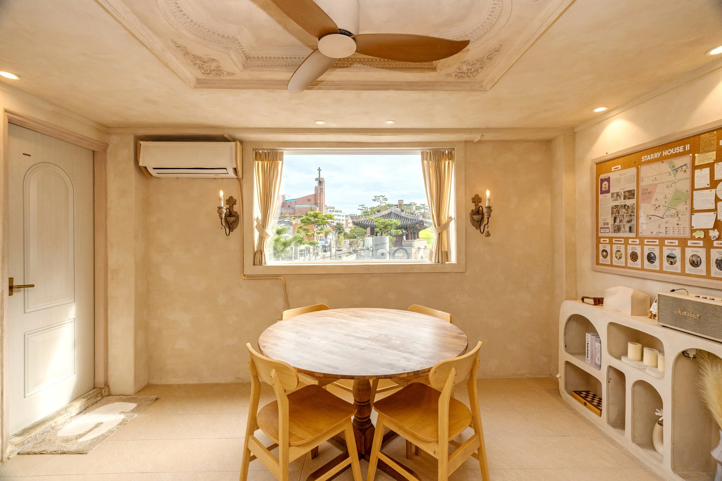 A cozy dining room with a round wooden table and four matching chairs, beige walls, a window with curtains showing a cityscape, a ceiling fan, wall-mounted lights, a bulletin board, and a white shelf with decorative items.