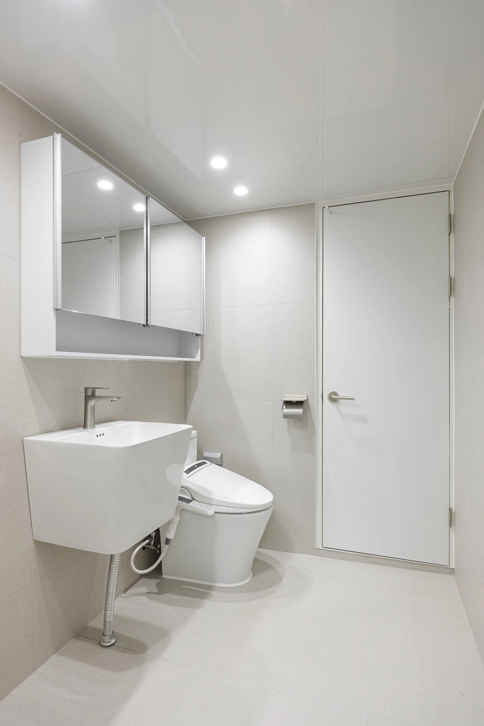 A modern, minimalist bathroom with a white toilet, a small sink with a faucet, a mirror cabinet overhead, a white door, a toilet paper holder, and recessed ceiling lights.