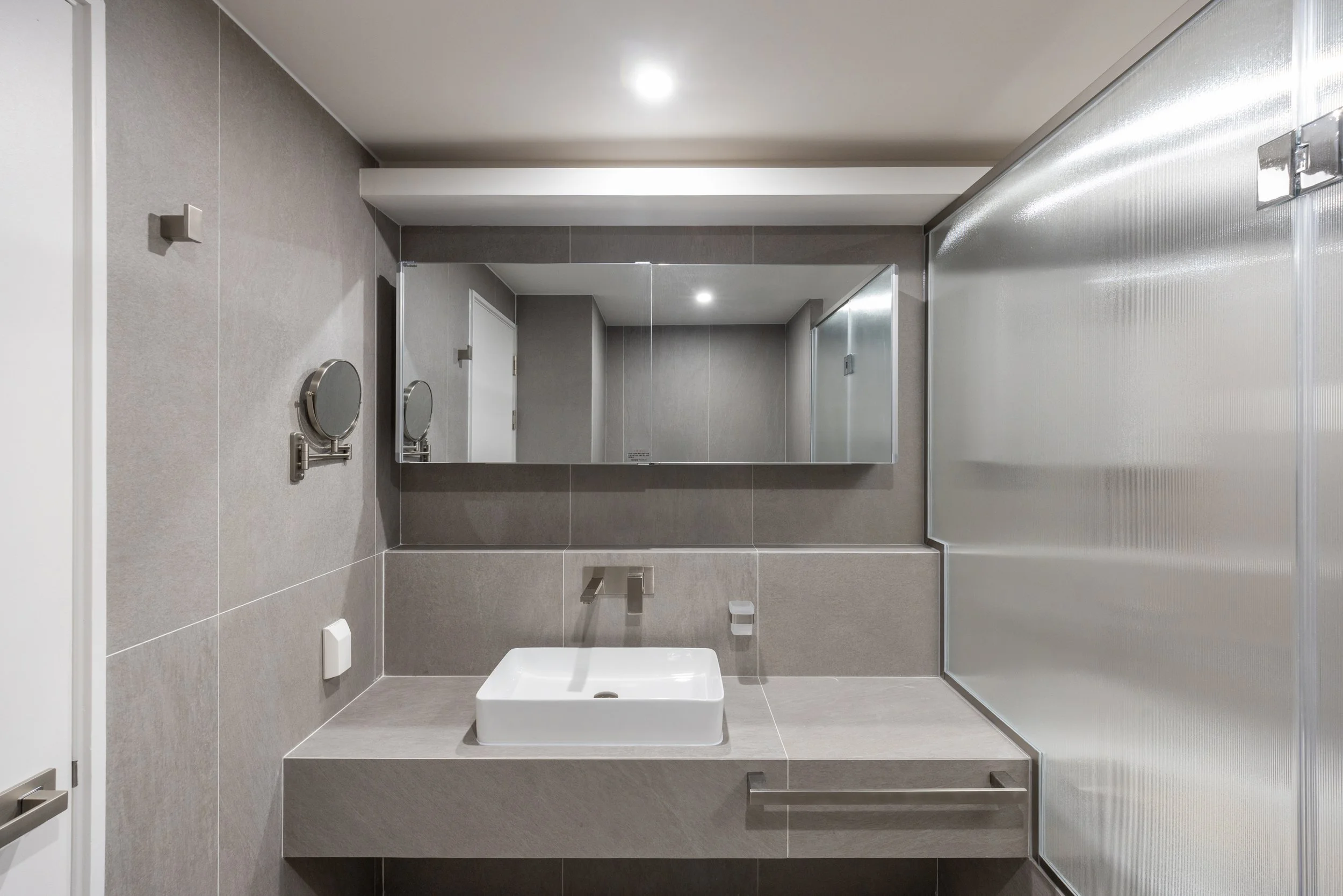 Modern bathroom with a white sink, gray tiled walls, a large mirror, and a metallic textured privacy divider.