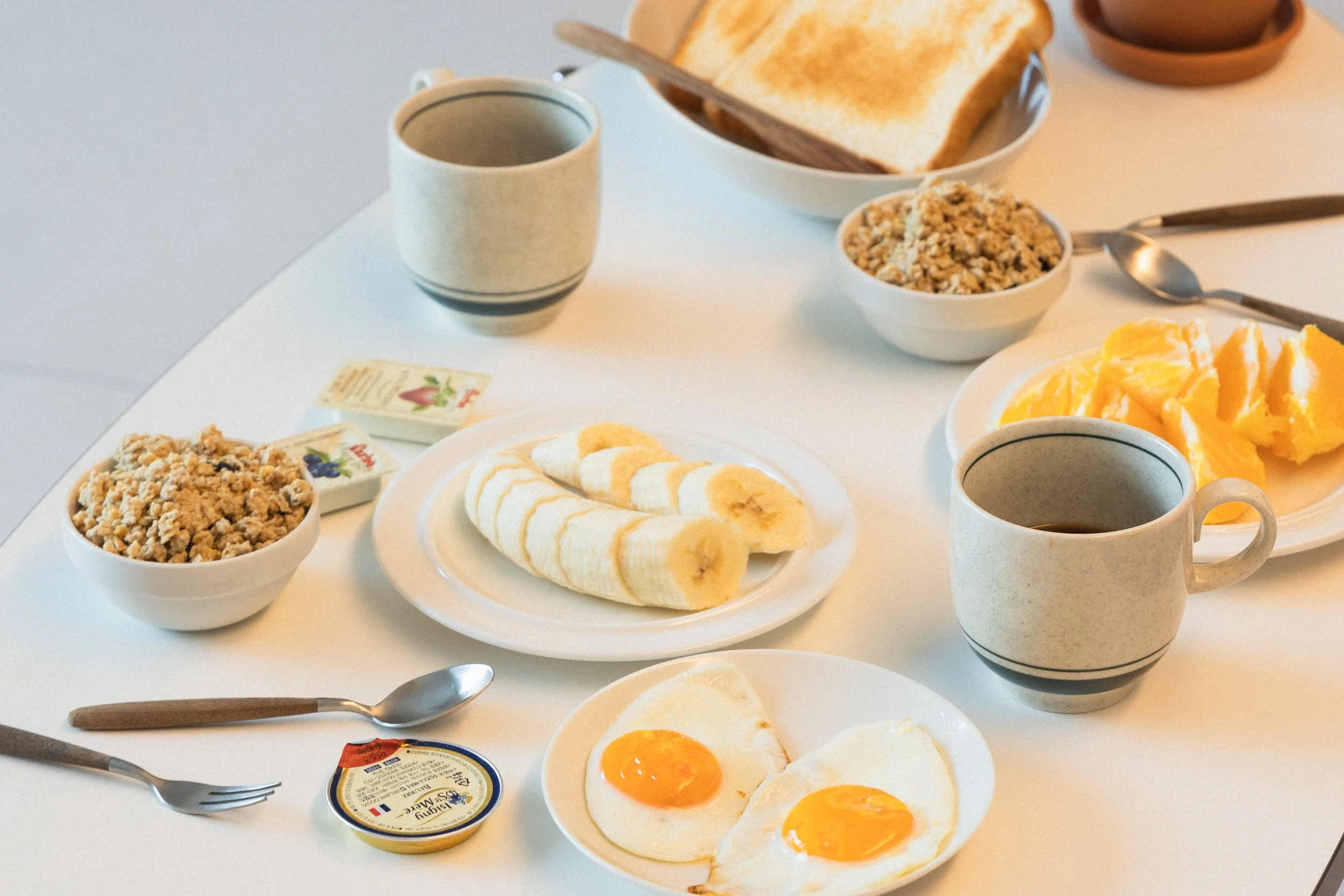 Breakfast spread with toast, banana slices, eggs, cereal, butter, jam, coffee, and butter on a white table.