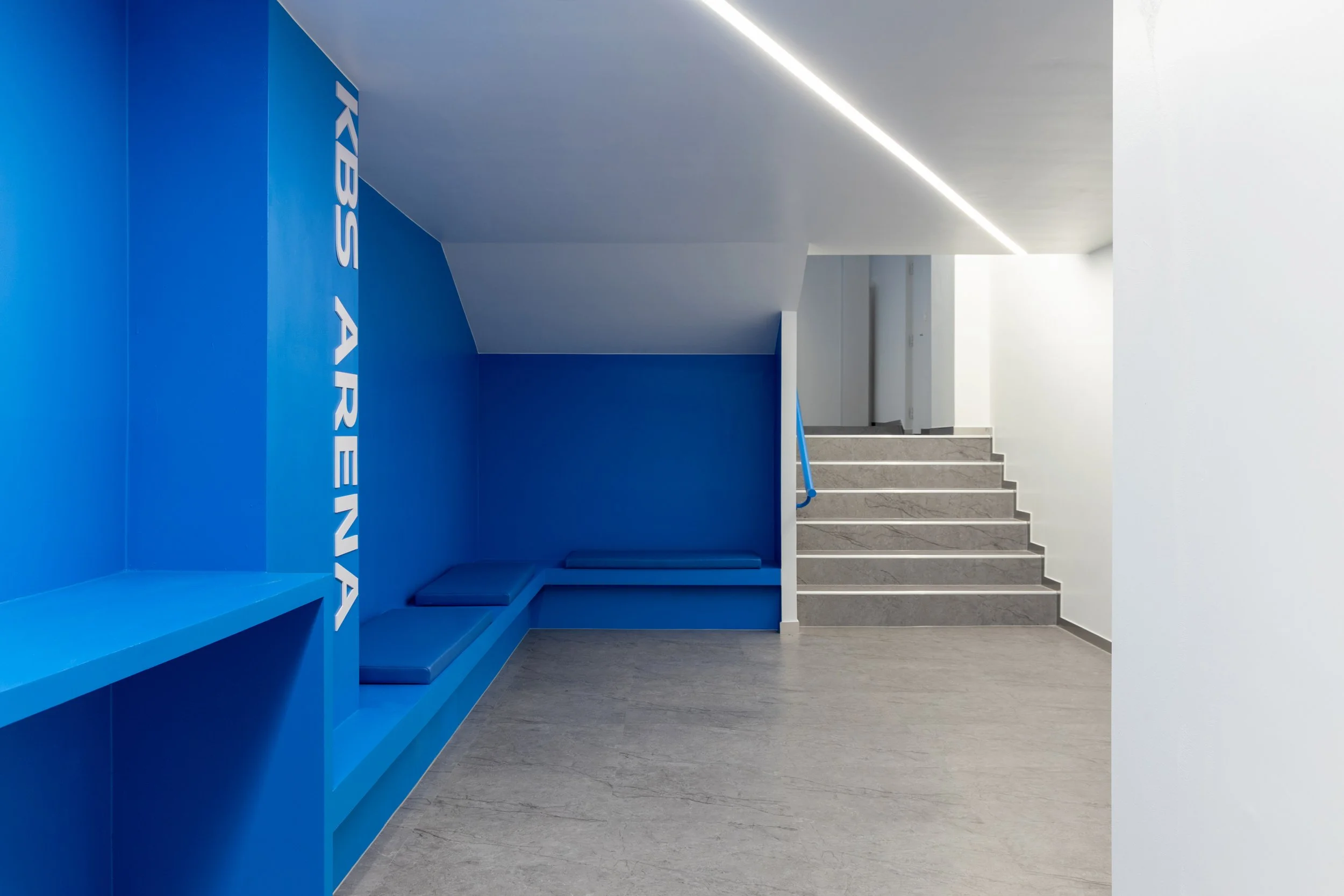 Modern interior with blue wall and seating area, stairs leading upward, minimalistic design, and natural light.