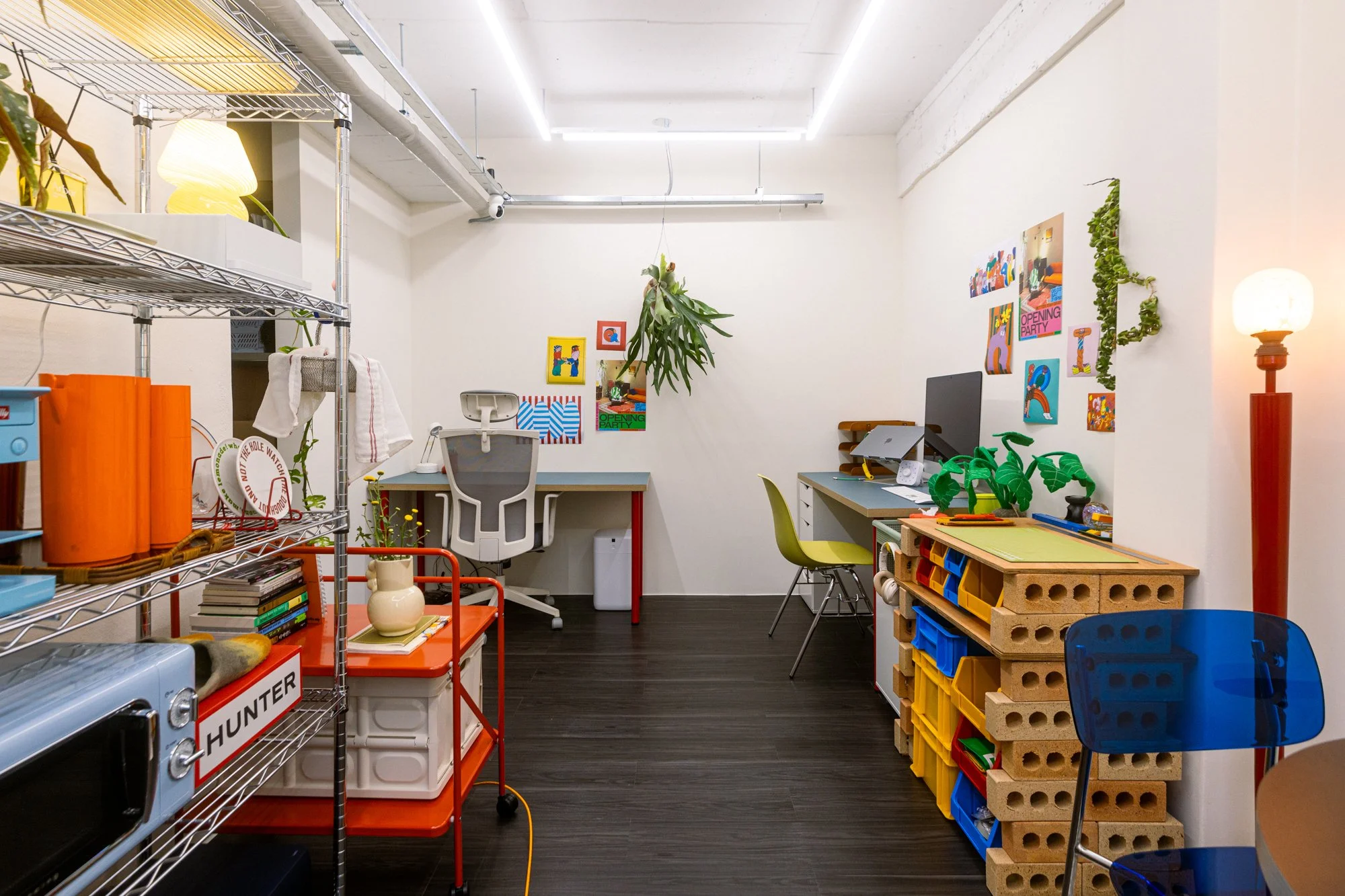 A colorful, organized workspace with shelves, desks, and decor, including a hanging plant, artwork on the wall, and a blue chair in the foreground.