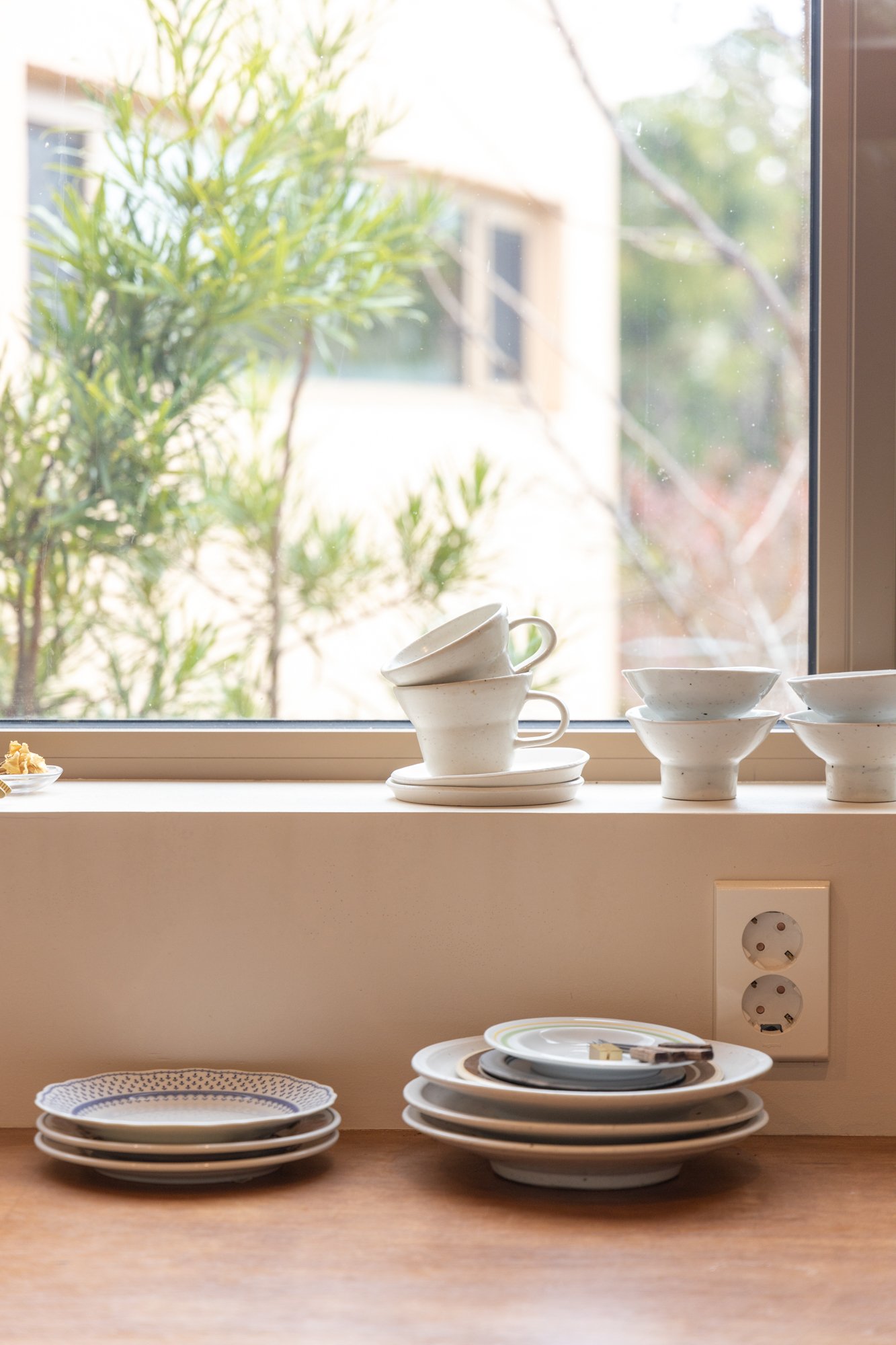 Stack of white ceramic bowls and plates on a wooden countertop near a window with a green outdoor view.