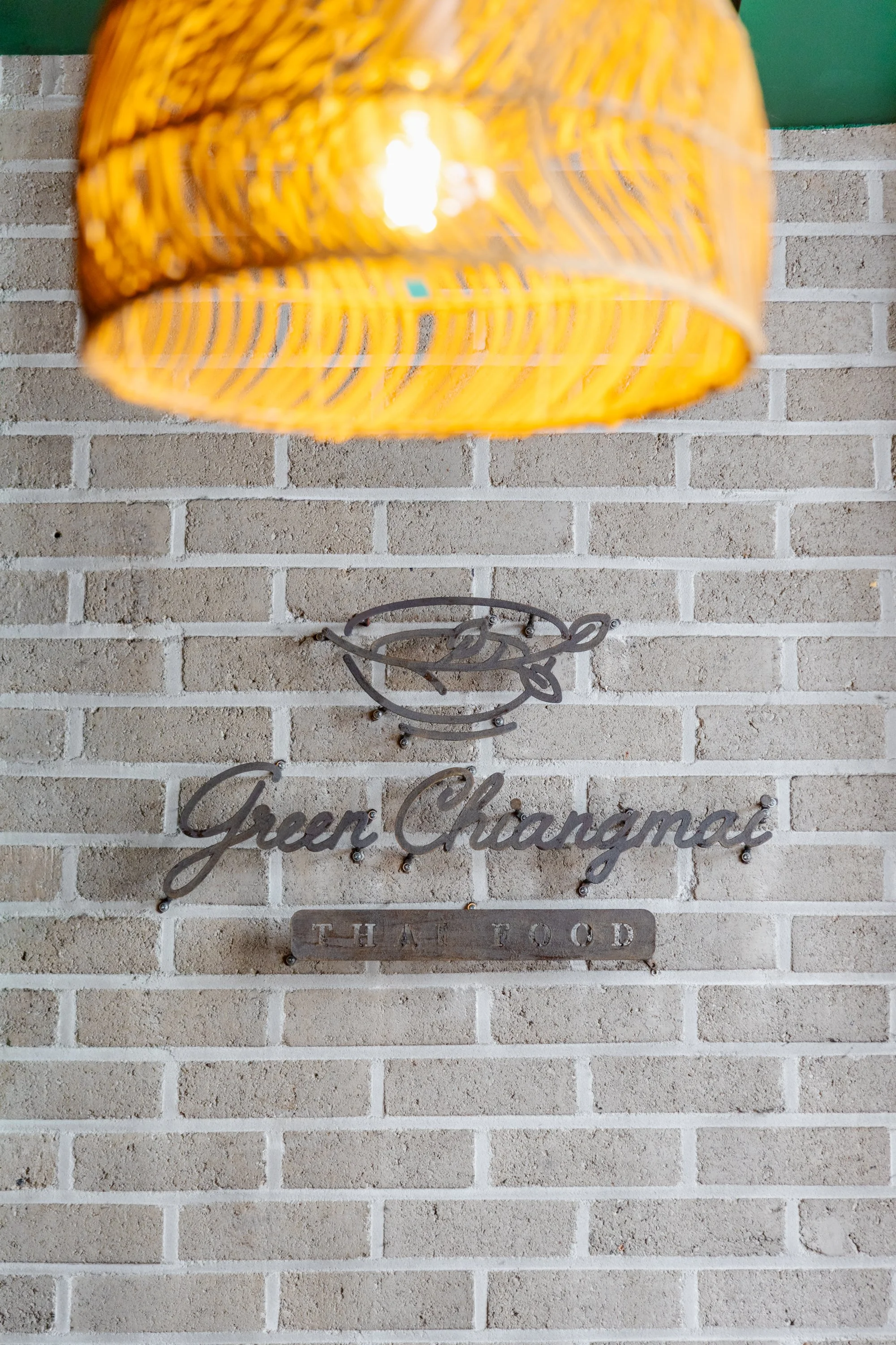 A wall with a sign that reads 'Green Changmai Thai Food,' featuring a fish logo above the text. Part of a yellow wicker hanging lamp is seen at the top of the image.