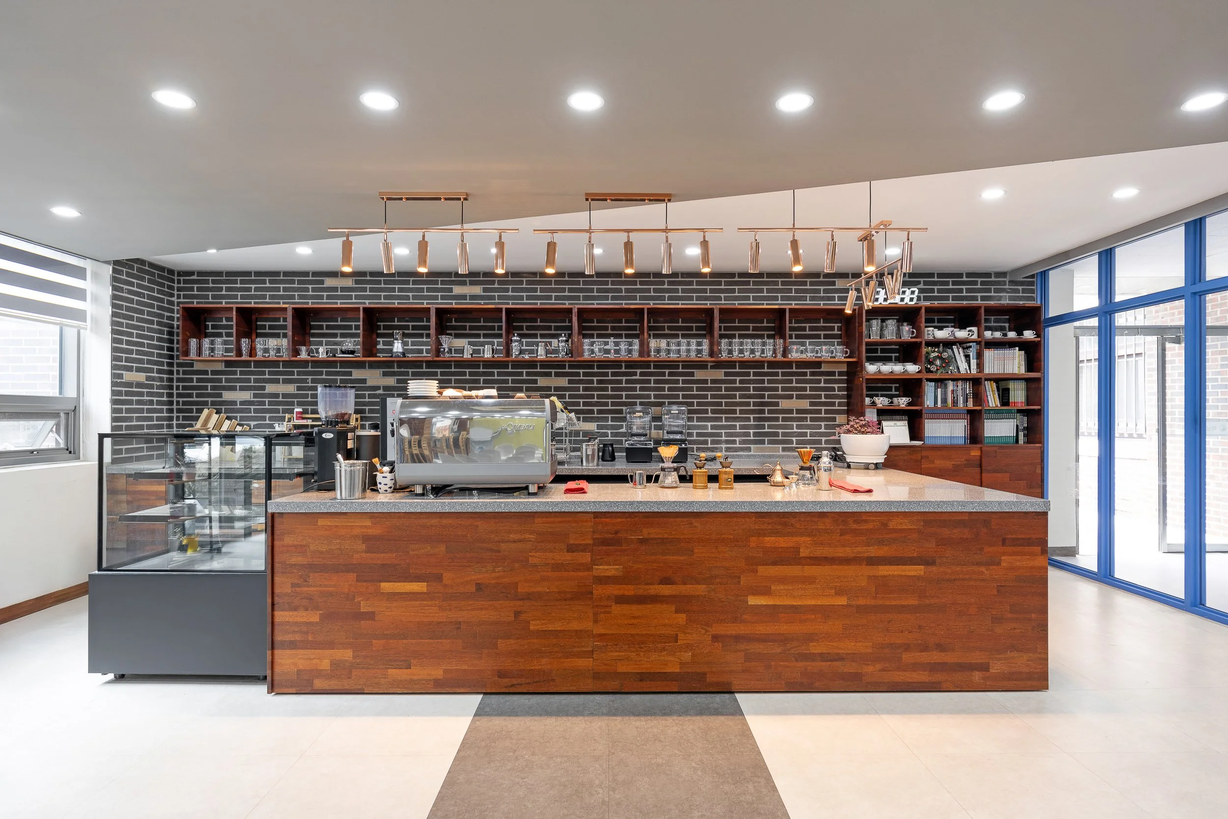 Modern coffee shop counter with espresso machine, coffee brewing equipment, glassware, and books on dark wood shelves, bright natural light from large windows.