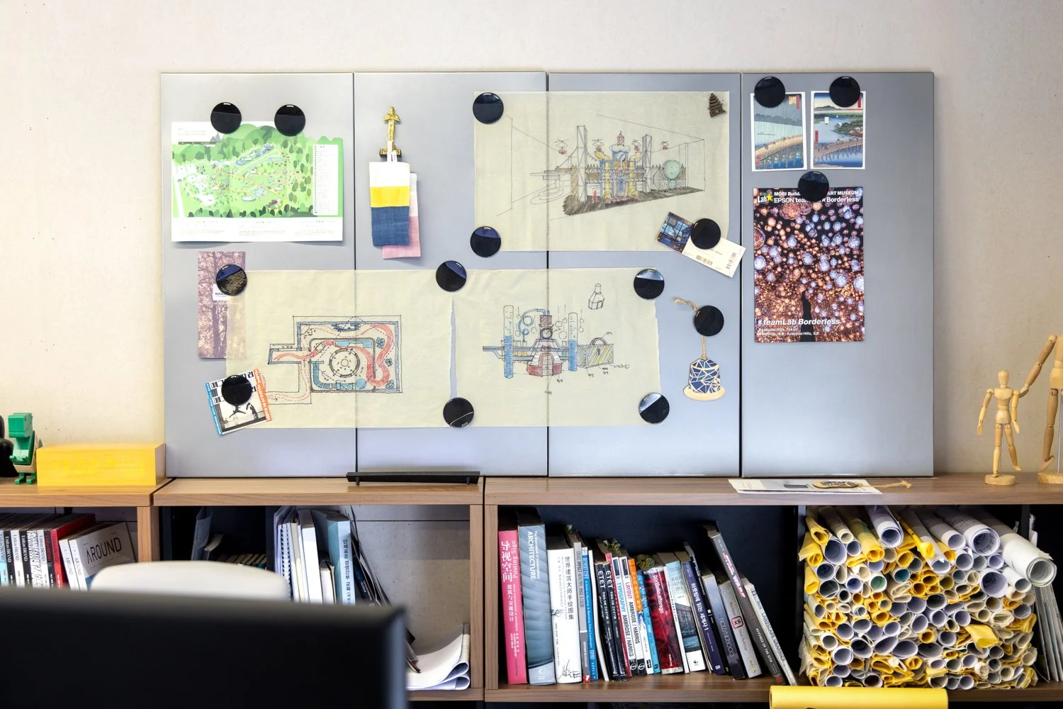 Organized workspace with a gray bulletin board filled with sketches, photos, and notes, above shelves containing rolled-up yellow and white papers, books, and a white chair.