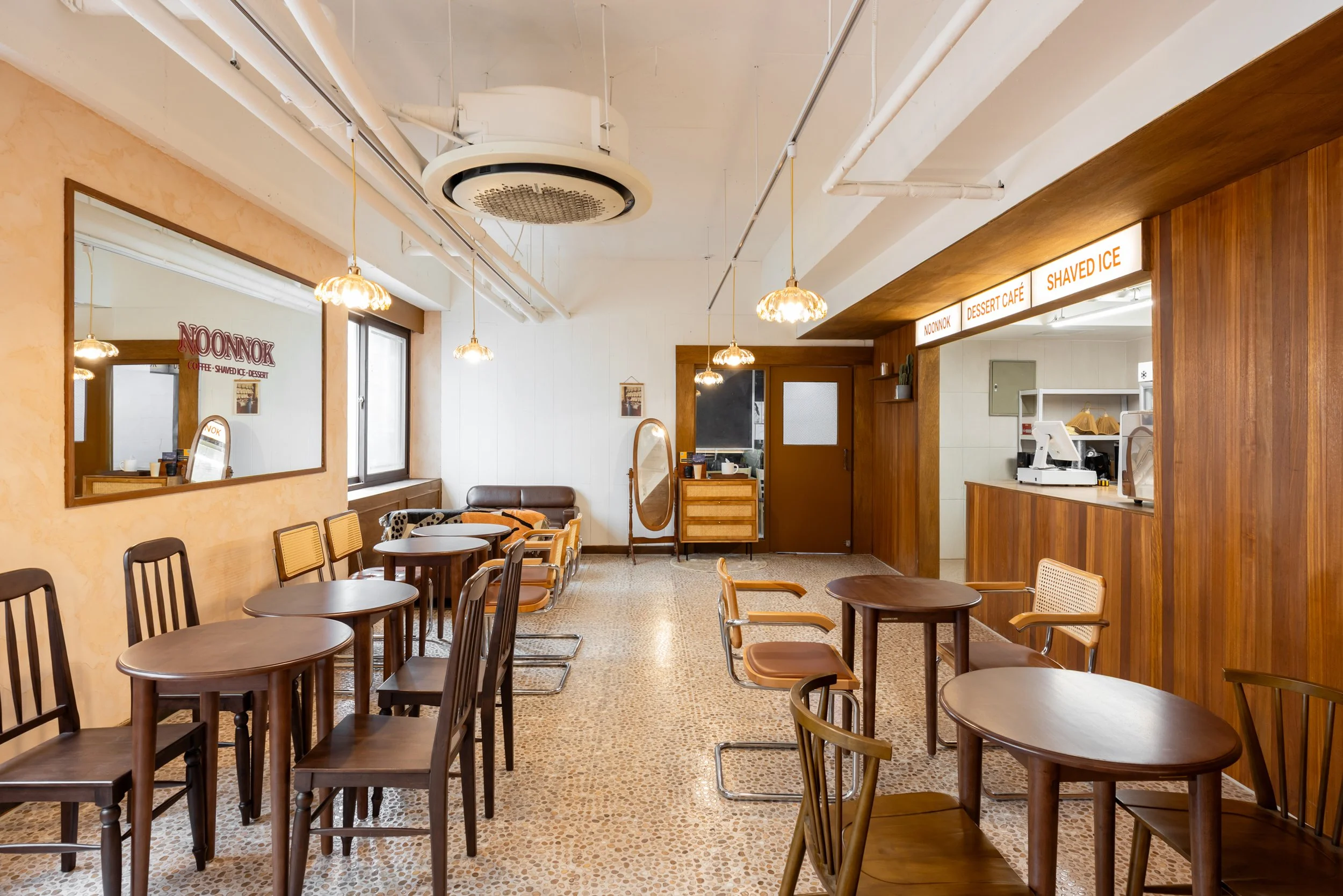 Empty cafe with wooden tables and chairs, a large window, mirror, and a counter area with a sign that says Noonnok, Dessert Cafe, Shaved Ice.